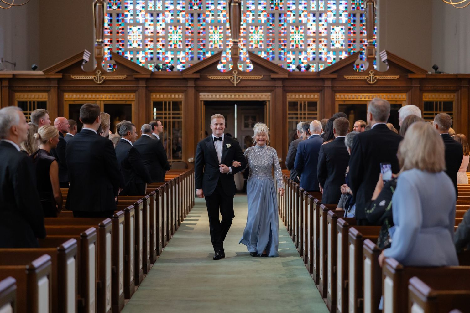 Elegant couple walking down church aisle with stained glass windows and wedding guests standing in pews.