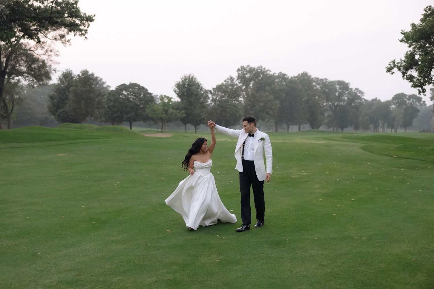 Bride and groom dance on a golf course lawn at dusk in elegant wedding attire.