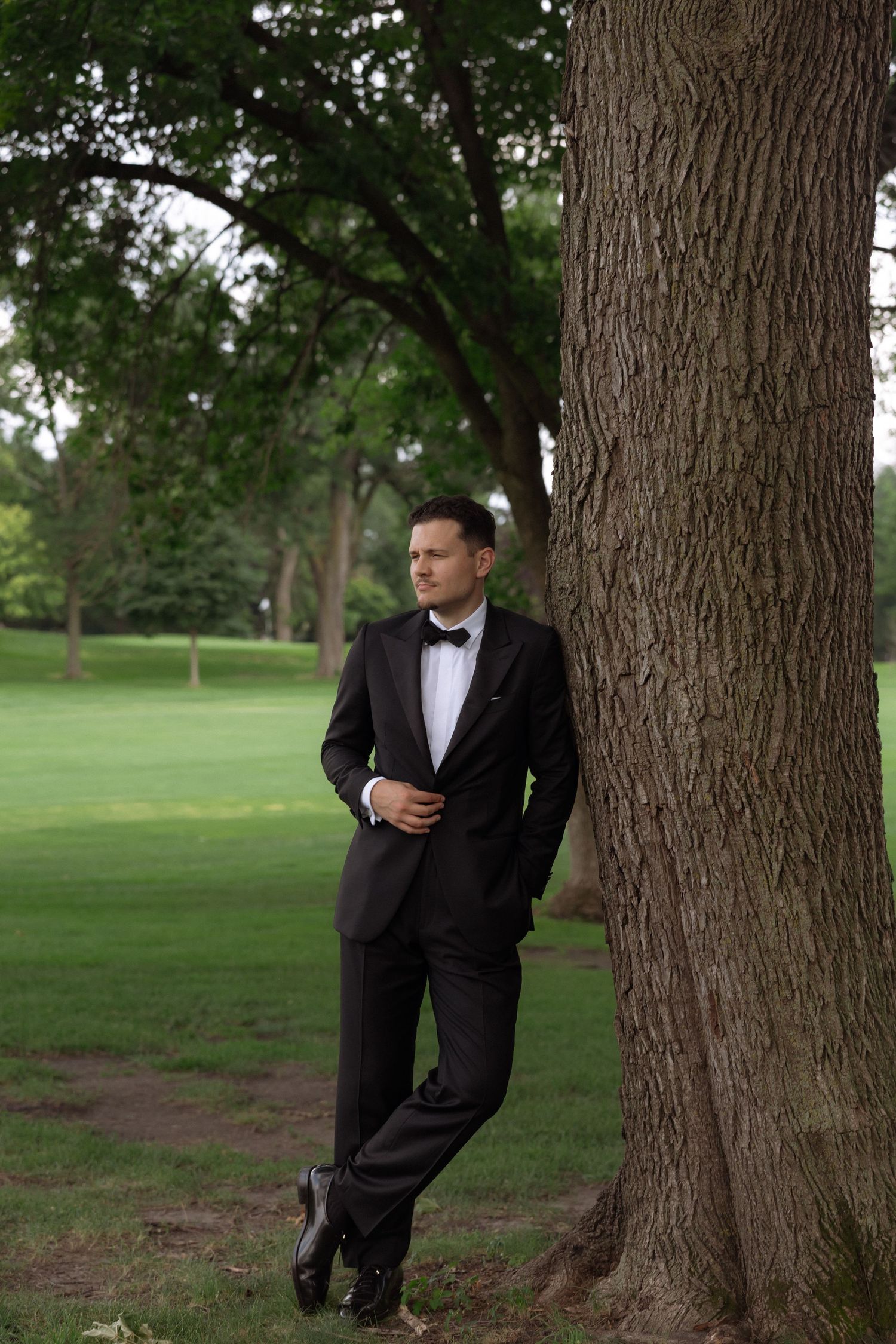 Dapper groom in black tuxedo leaning against large tree trunk on lush green golf course.