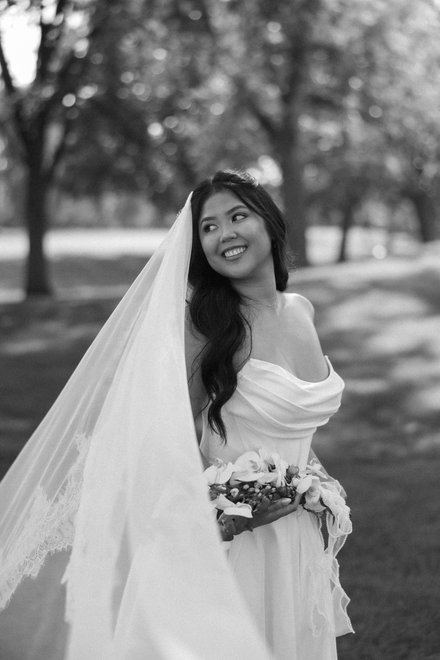 Smiling Asian bride in off-shoulder gown holds bouquet with veil flowing in black and white.