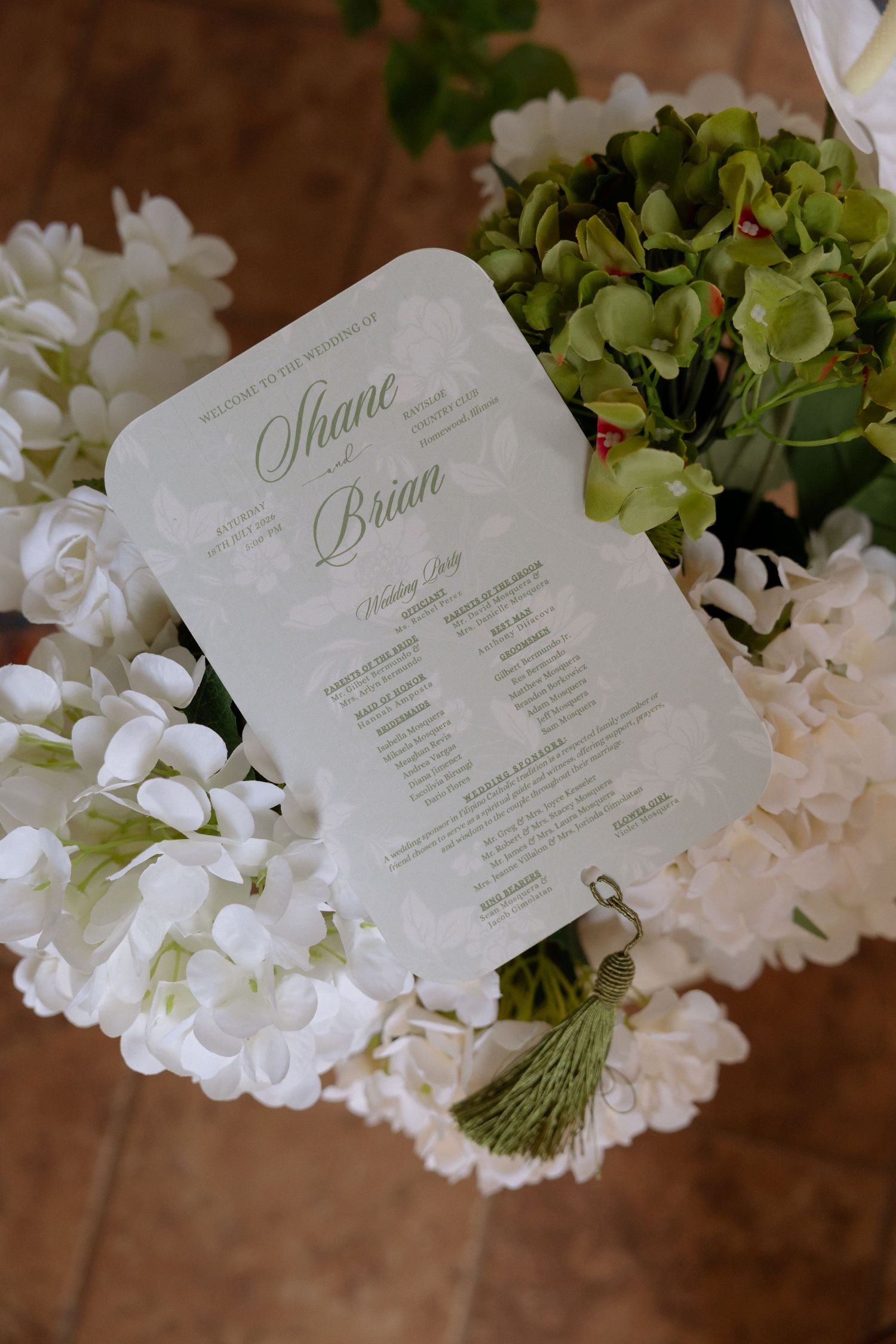 Elegant wedding program for Shane and Brian displayed among white hydrangeas and green floral bouquet with tassel detail.