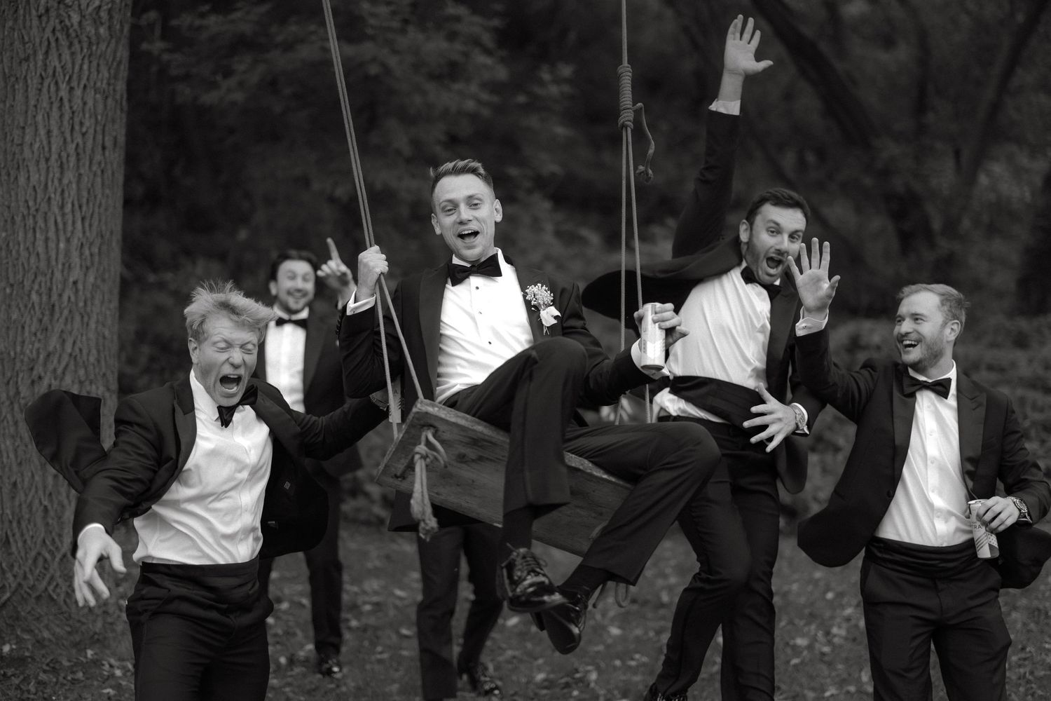 Groom on swing surrounded by laughing groomsmen in tuxedos during fun black and white wedding photo.
