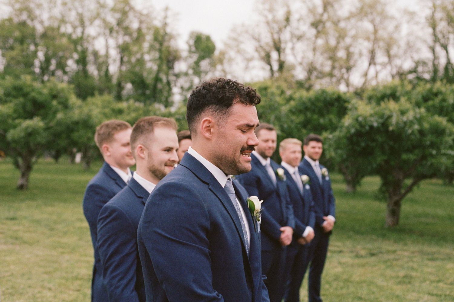 Groom and groomsmen in navy suits standing outdoors among trees at a wedding ceremony.