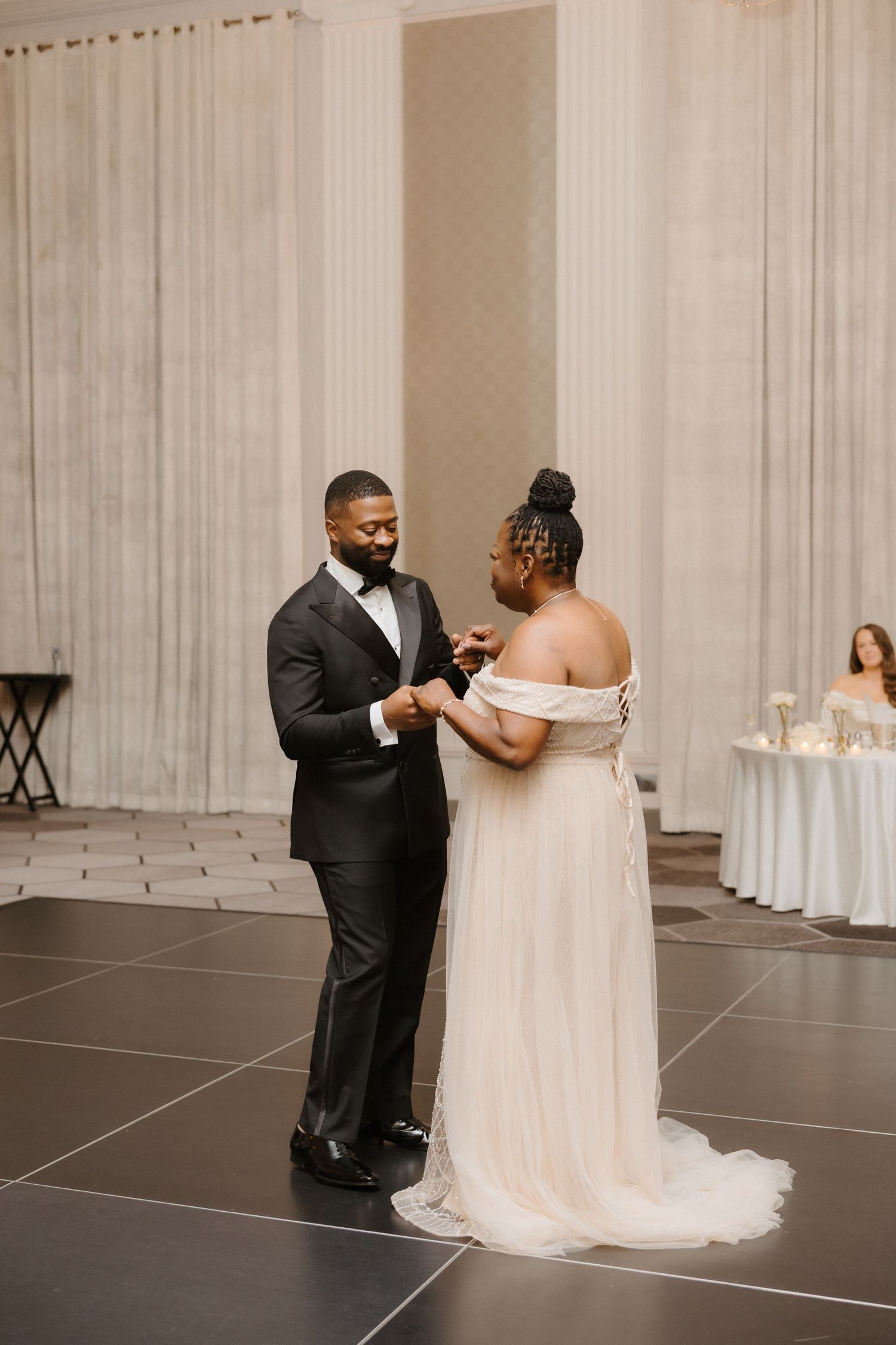 A couple shares their first dance at an elegant wedding reception, groom in a black tuxedo and bride in an off-shoulder white gown.