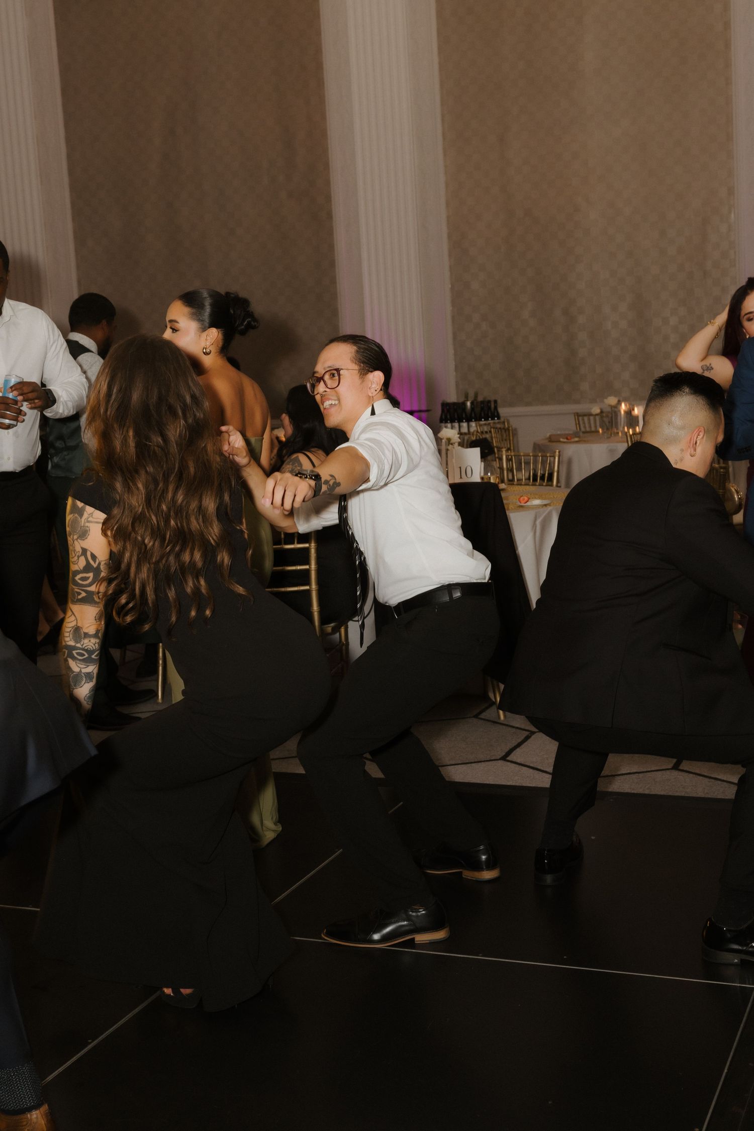 Guests dancing energetically at a wedding reception in an elegant ballroom with gold chairs and decorative columns.
