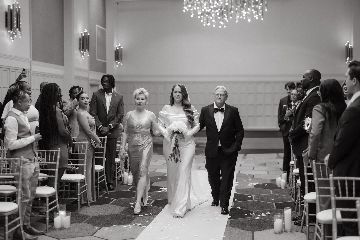 Bride walks down the aisle with parents in elegant black and white wedding ceremony in a grand ballroom.