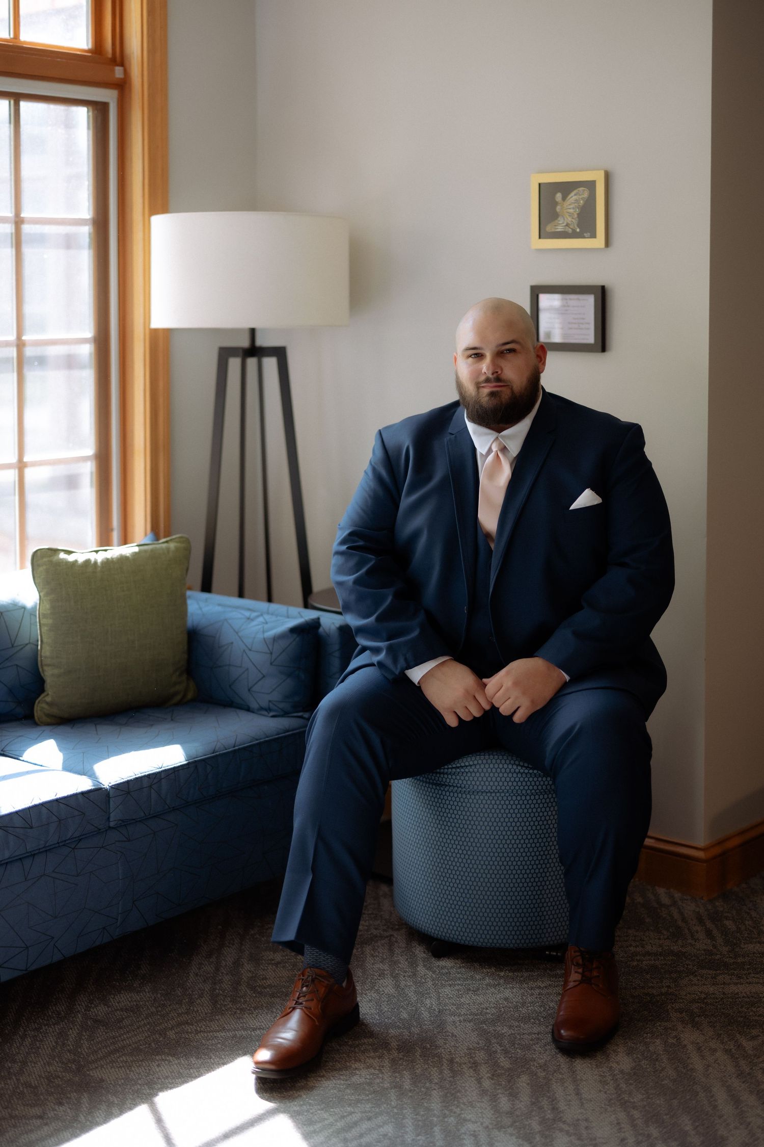 A bald bearded man in a navy suit with pink tie sits confidently on a ottoman in an elegant room with natural light.