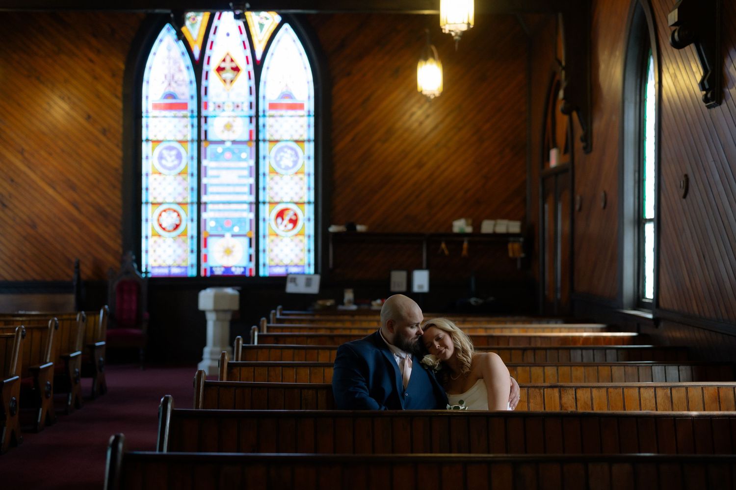 Couple sitting closely together in wooden church pews beneath beautiful stained glass windows during intimate wedding ceremony.