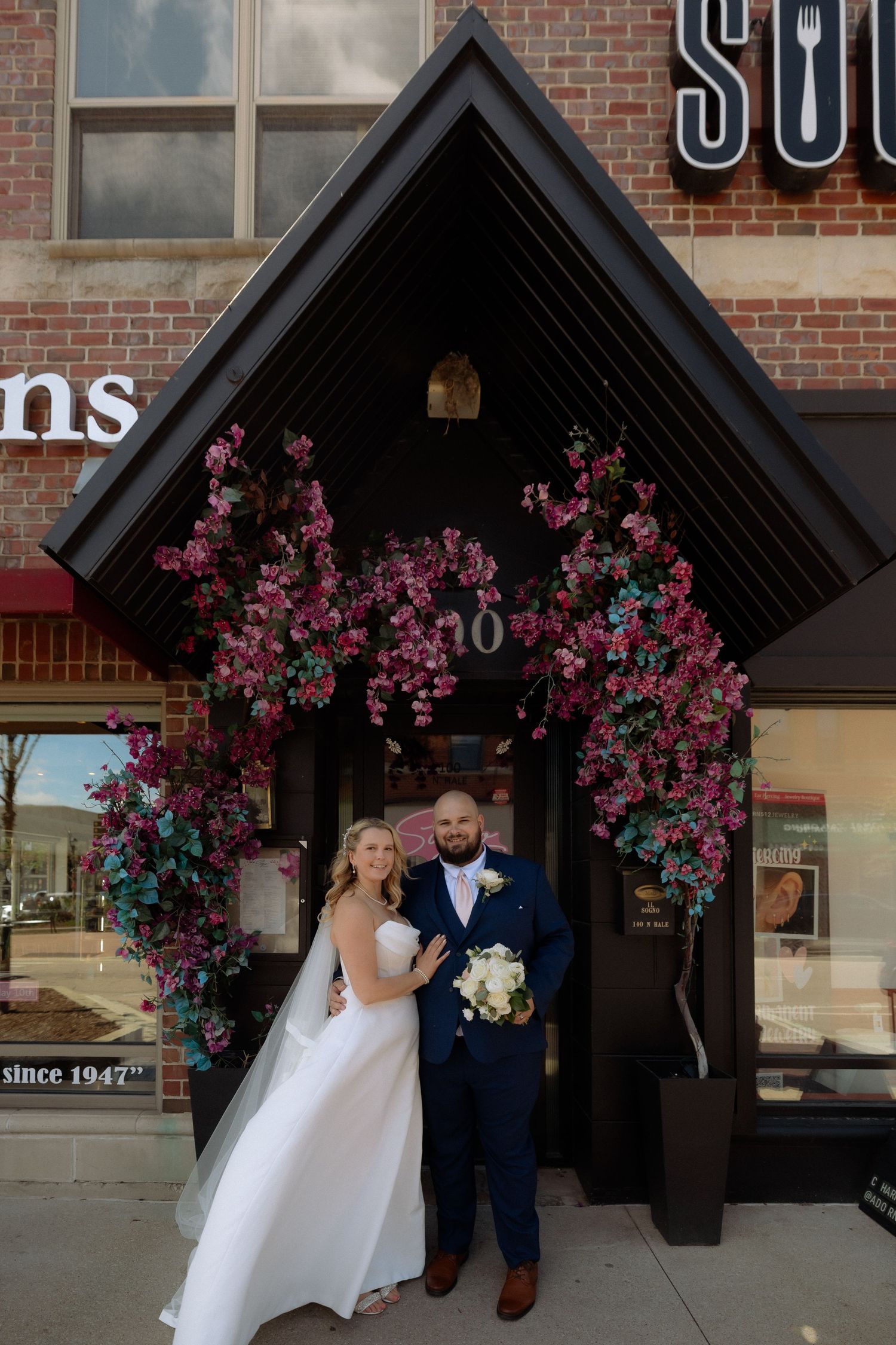 Bride and groom posing under a floral arch with pink blooms at a brick building entrance on their wedding day.