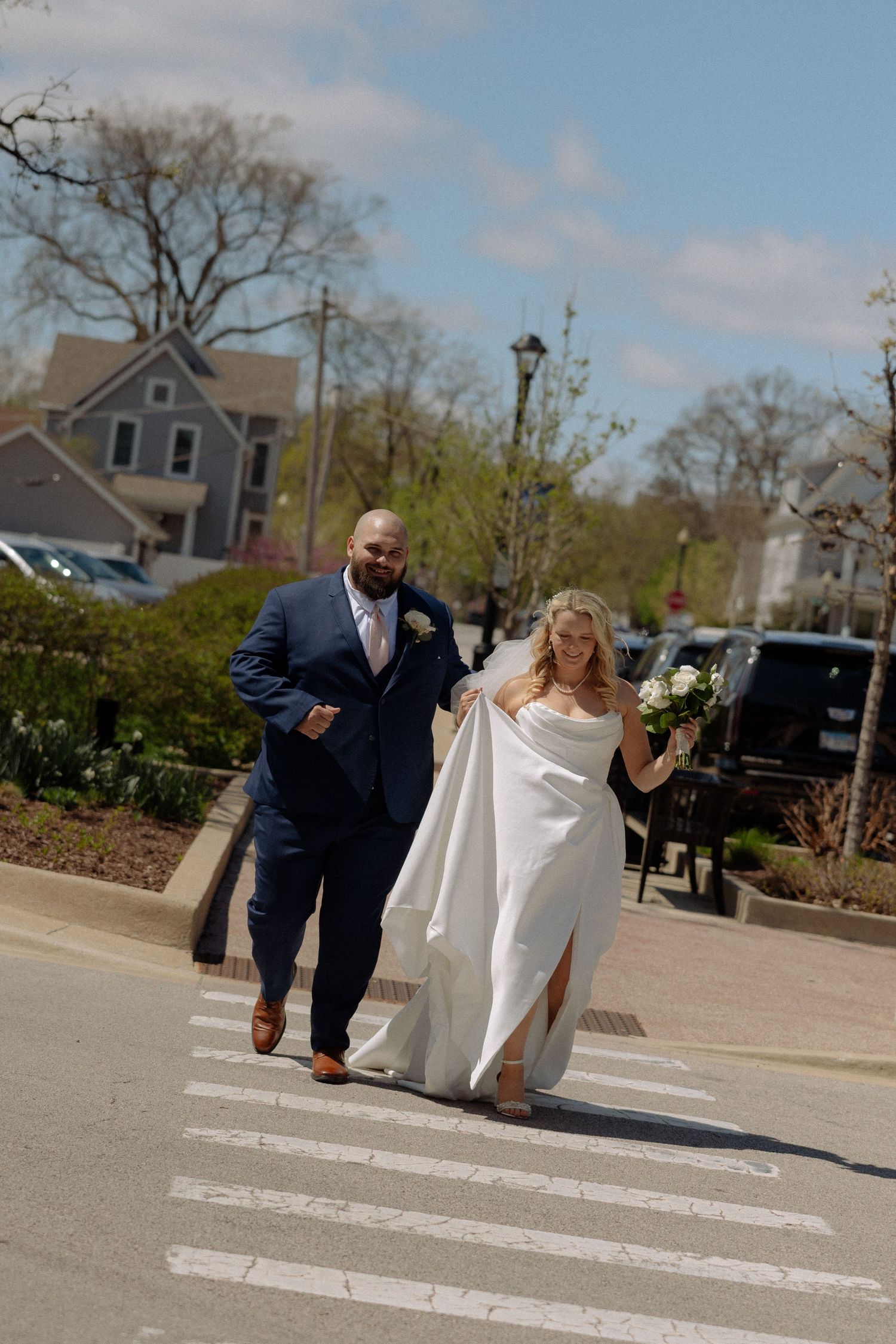 A bride and groom cross a sunny street in a charming town, she in a white gown holding flowers, he in a navy suit.