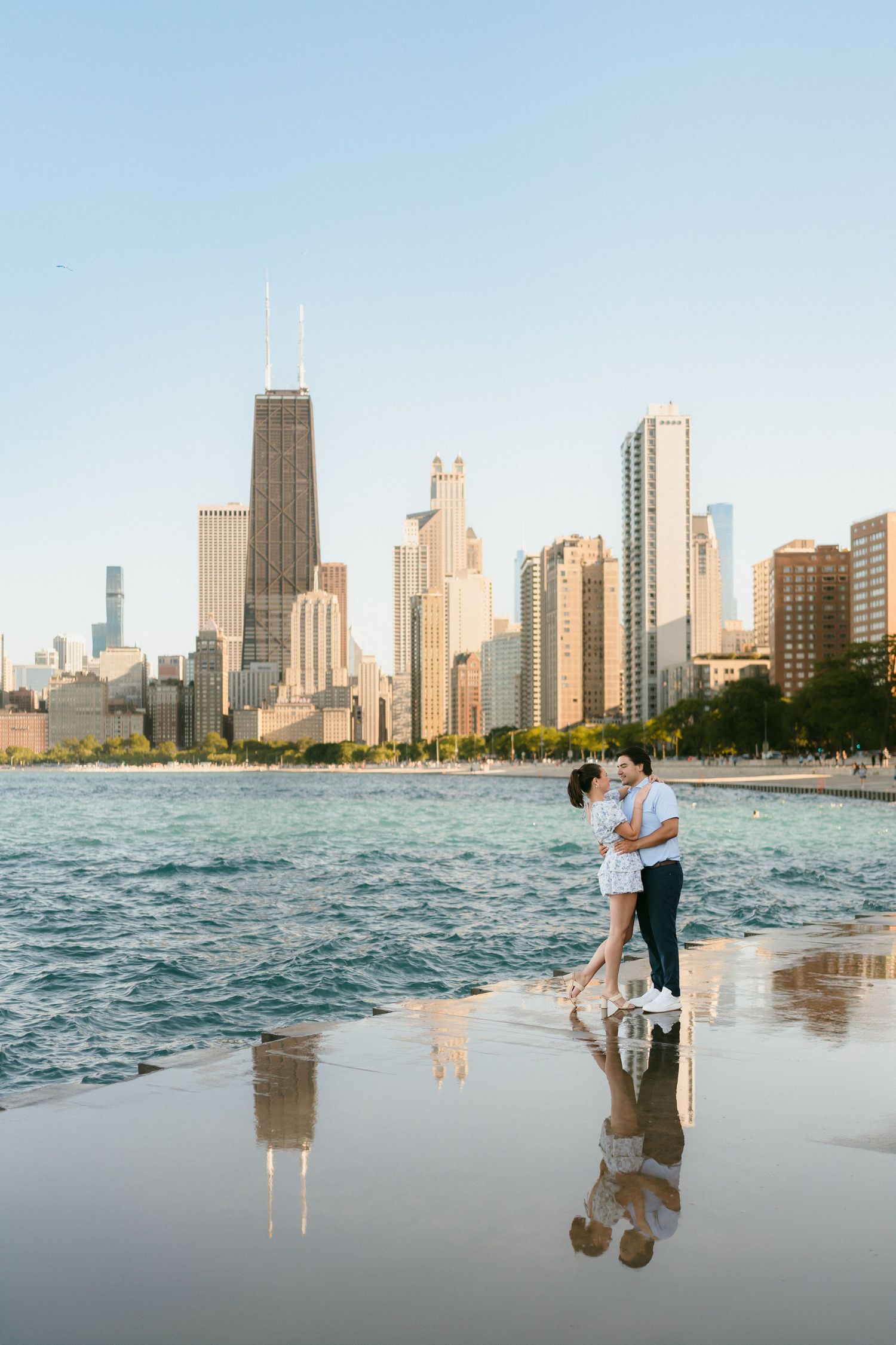 Romantic couple kissing on wet beach sand with stunning Chicago skyline reflected in water at golden hour.