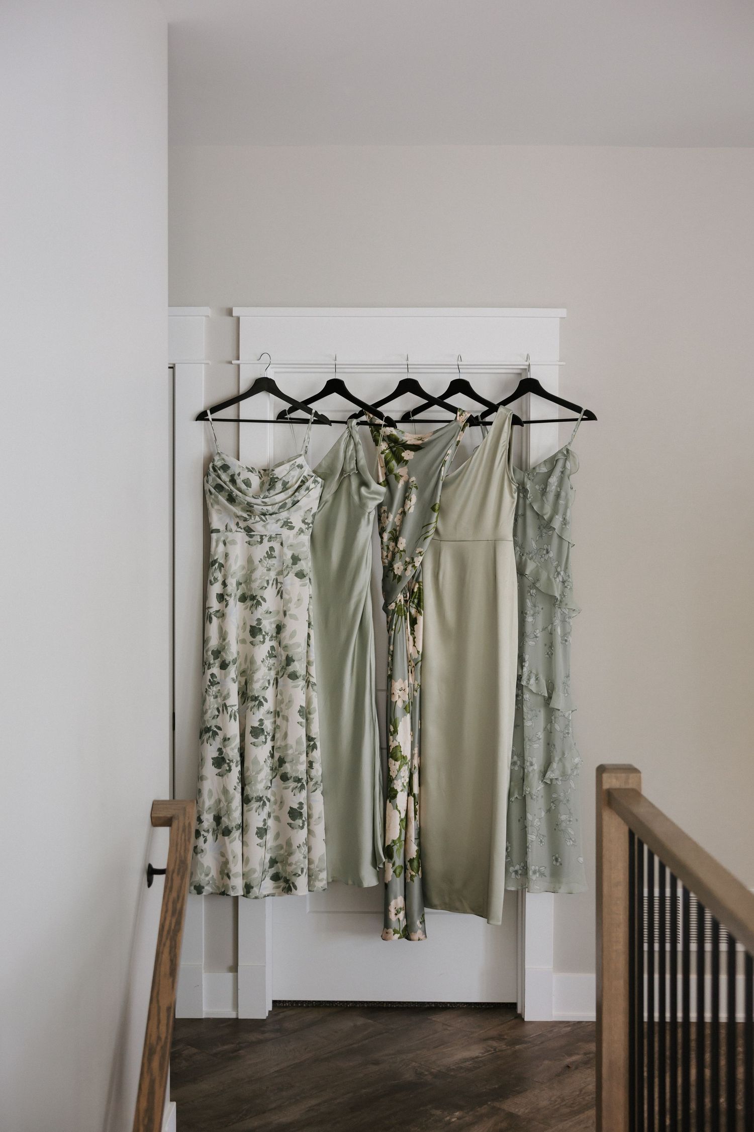 Four sage green and floral bridesmaid dresses hanging on black hangers at a wedding venue.
