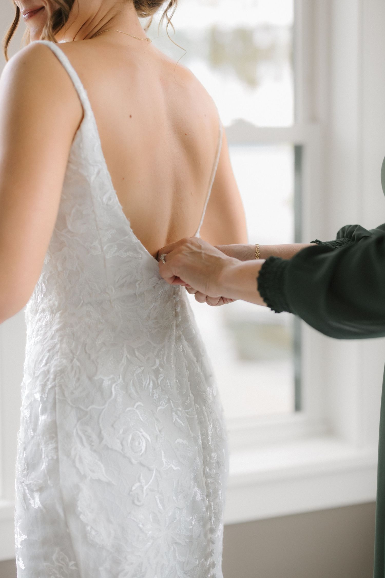 Close-up of hands fastening buttons on back of bride's white lace low-back wedding dress by window.