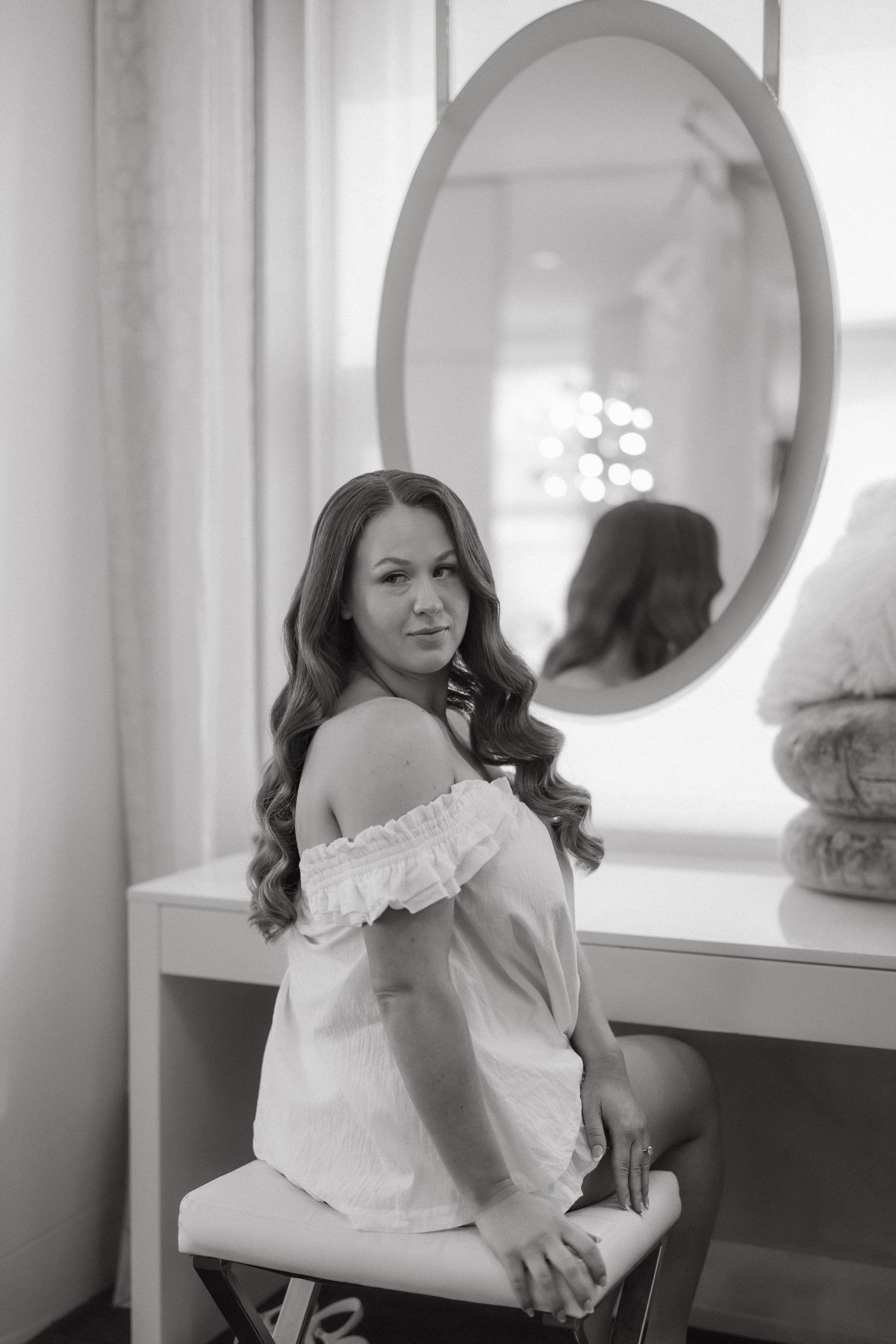 Black and white photo of a woman with long wavy hair sitting at a vanity table, wearing an off-shoulder top.