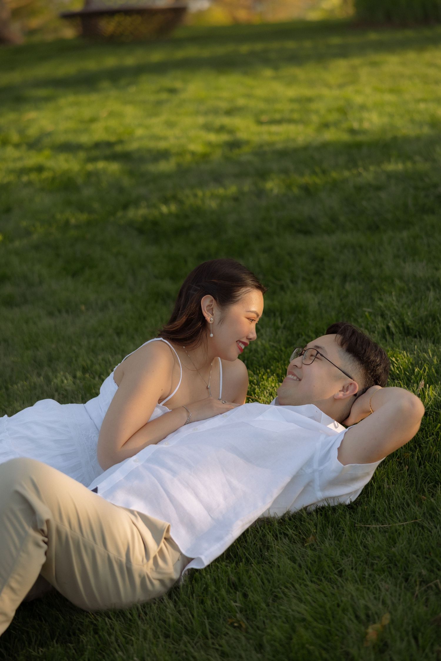 Couple relaxing on green grass in golden hour light, woman in white dress smiling at man lying down.