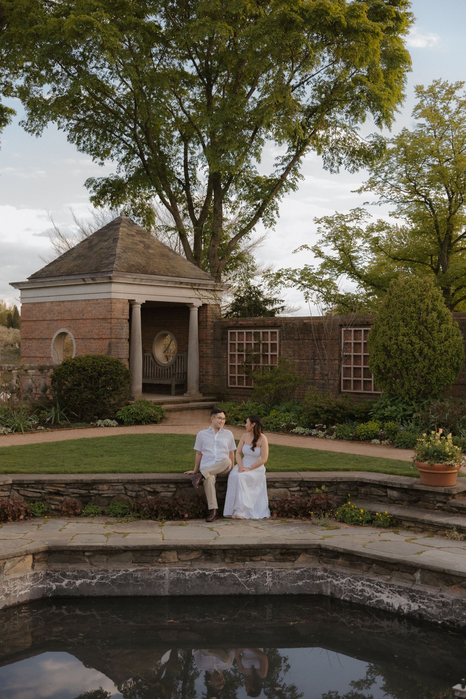 Couple in white attire sitting by a reflecting pool in a formal garden with a brick pavilion.
