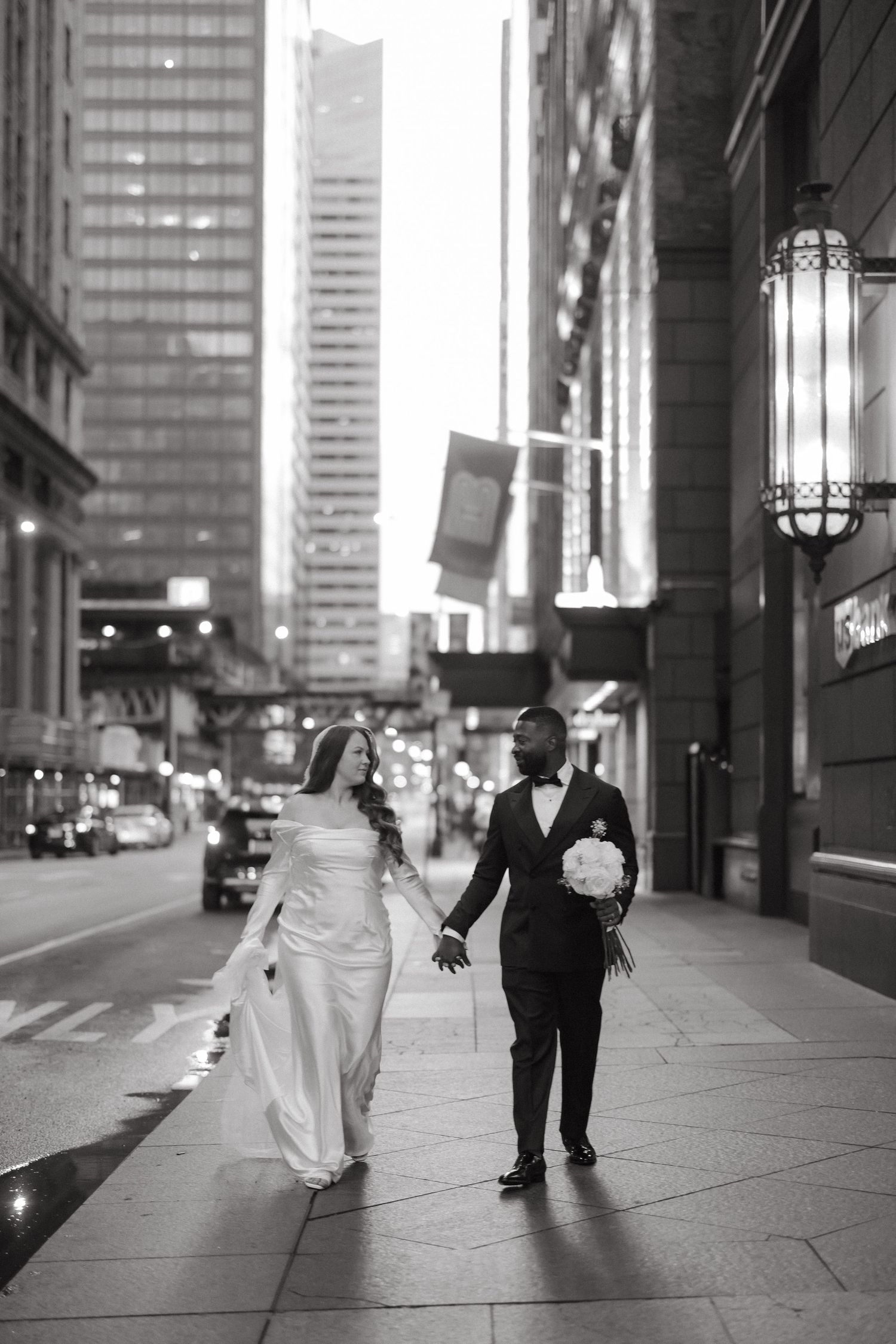 Black and white photo of a bride and groom holding hands walking on a city sidewalk in wedding attire.