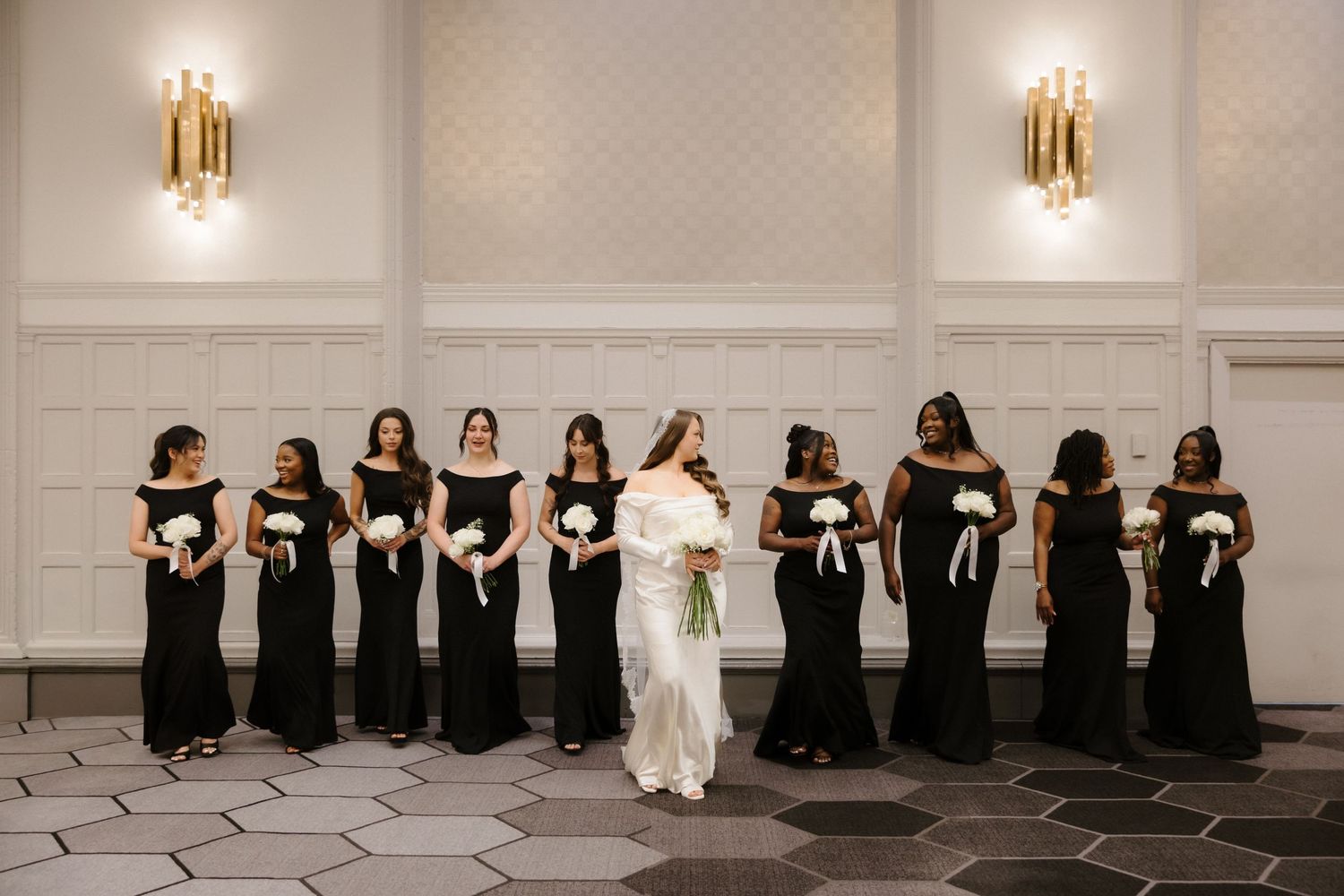 Bride in white gown poses with bridesmaids in black off-shoulder dresses holding white bouquets in elegant ballroom.