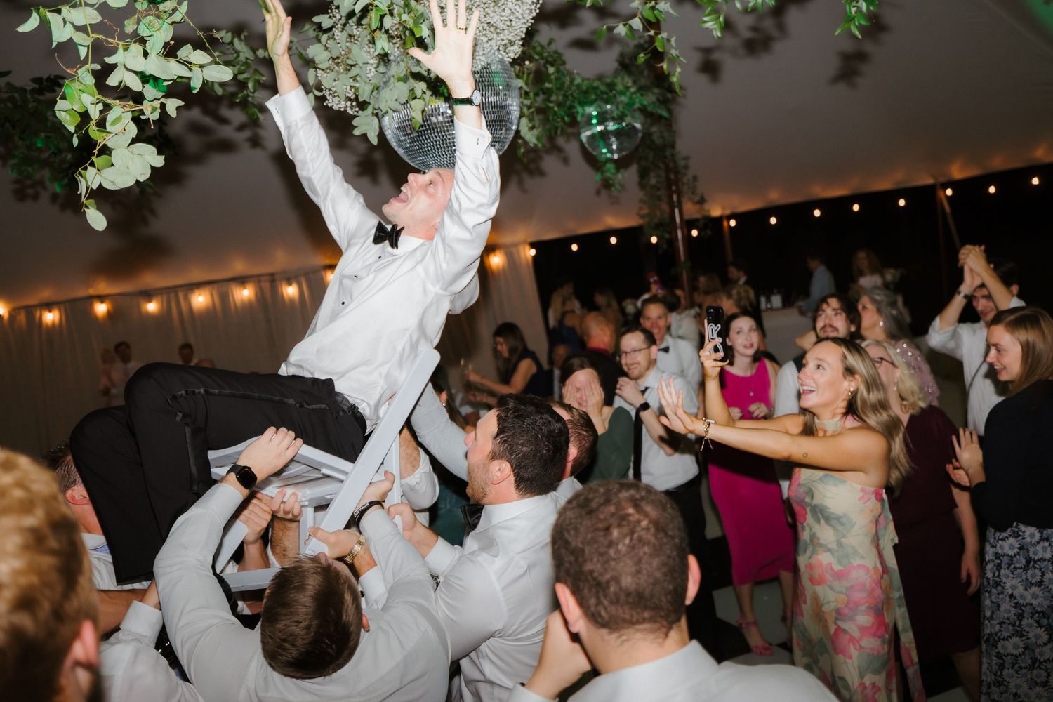 Groom is lifted by groomsmen on crowded wedding dance floor under disco ball and floral decorations.