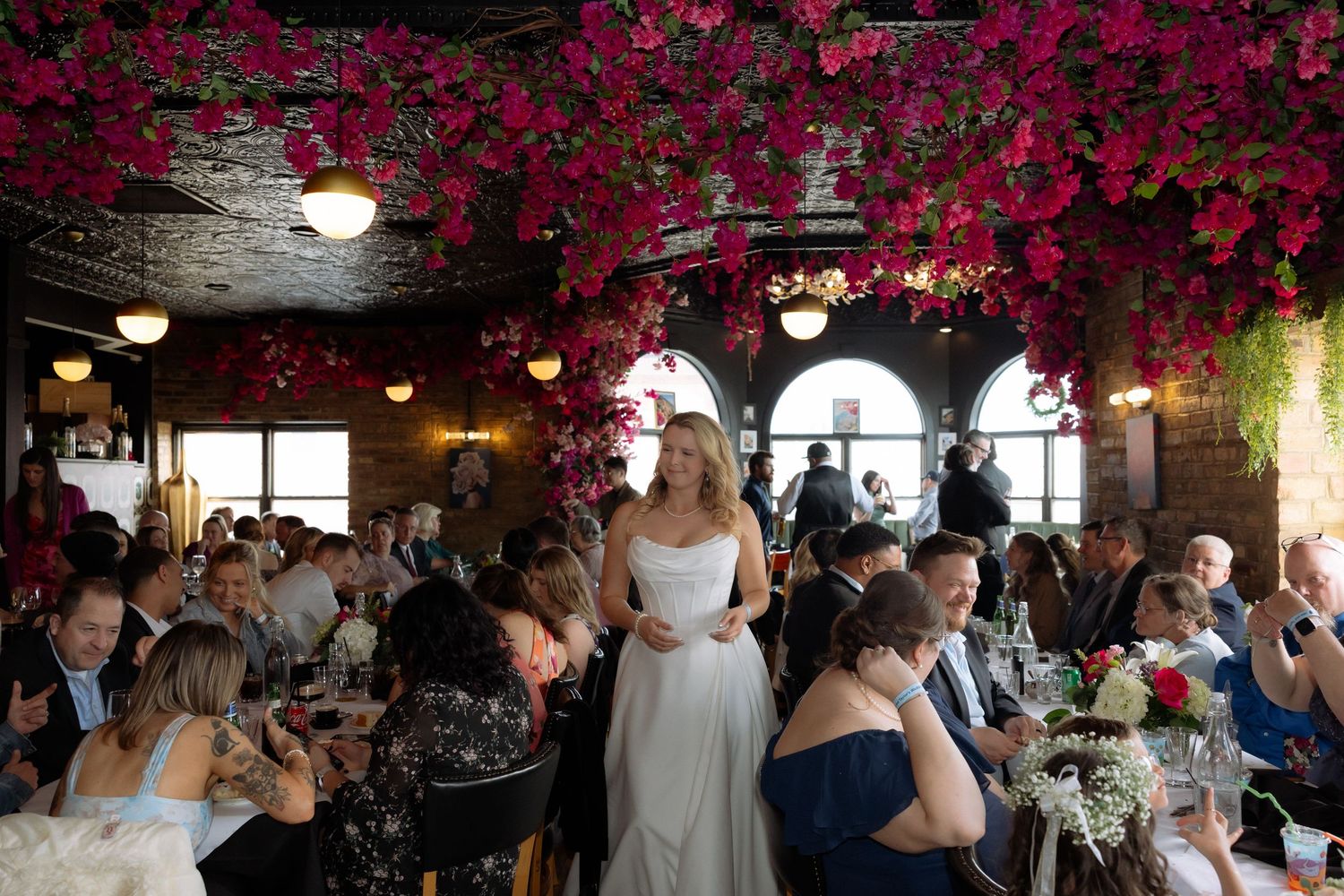 Bride in white gown stands among wedding guests under stunning pink floral ceiling installation in elegant venue.
