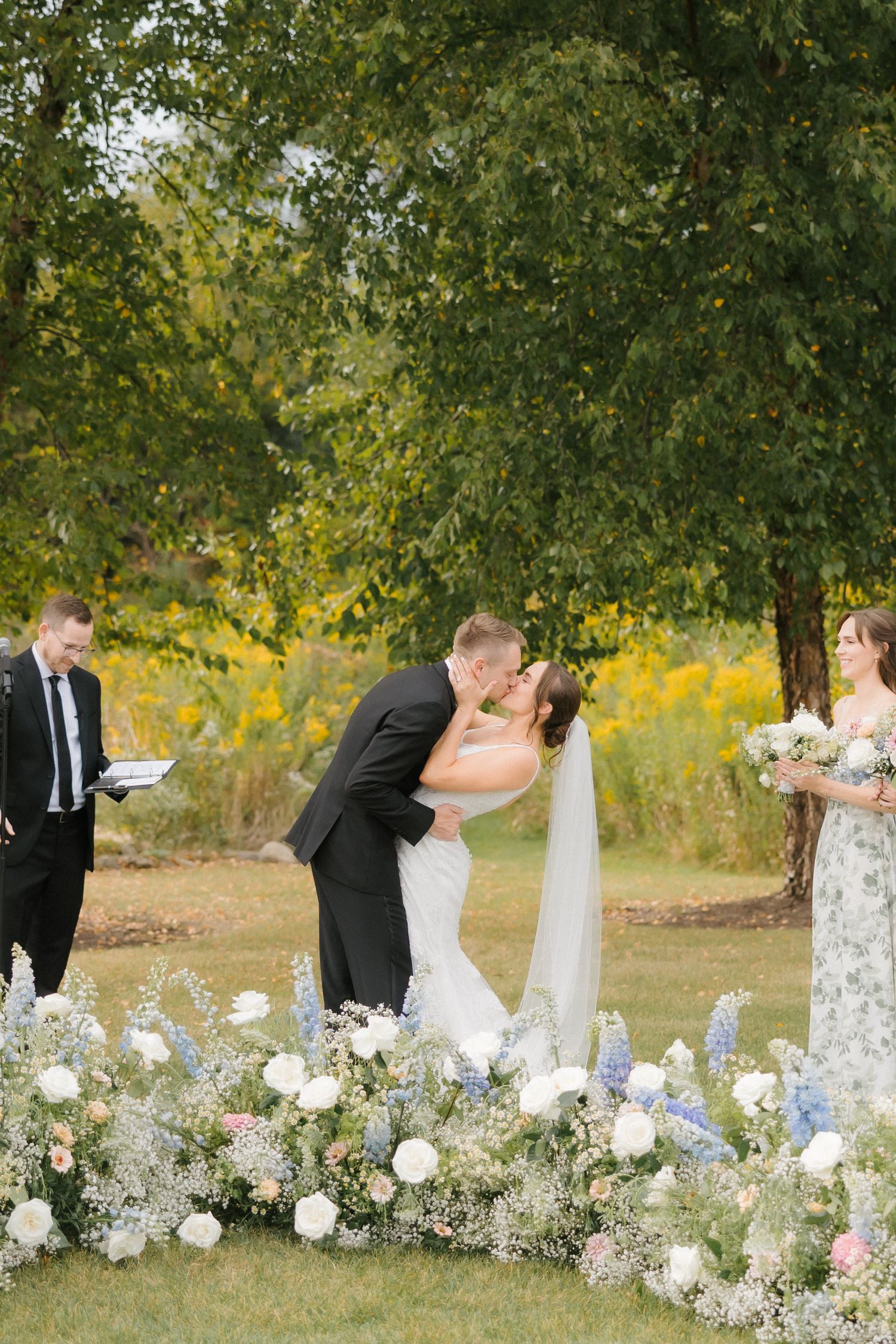 Bride and groom share first kiss at outdoor ceremony surrounded by lush floral ground arrangements.