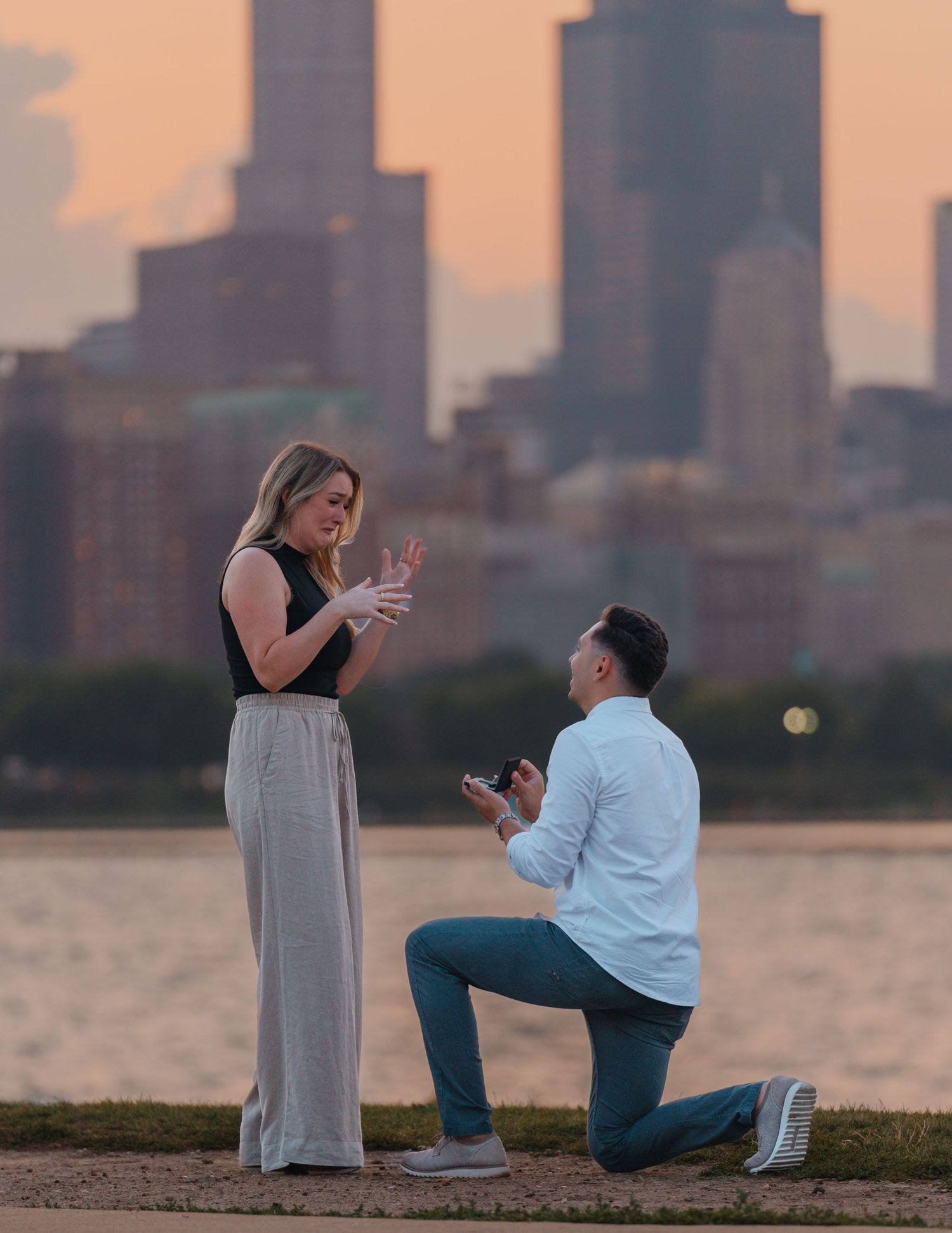 Emotional woman reacting to a marriage proposal as her partner kneels with a ring by Lake Michigan.