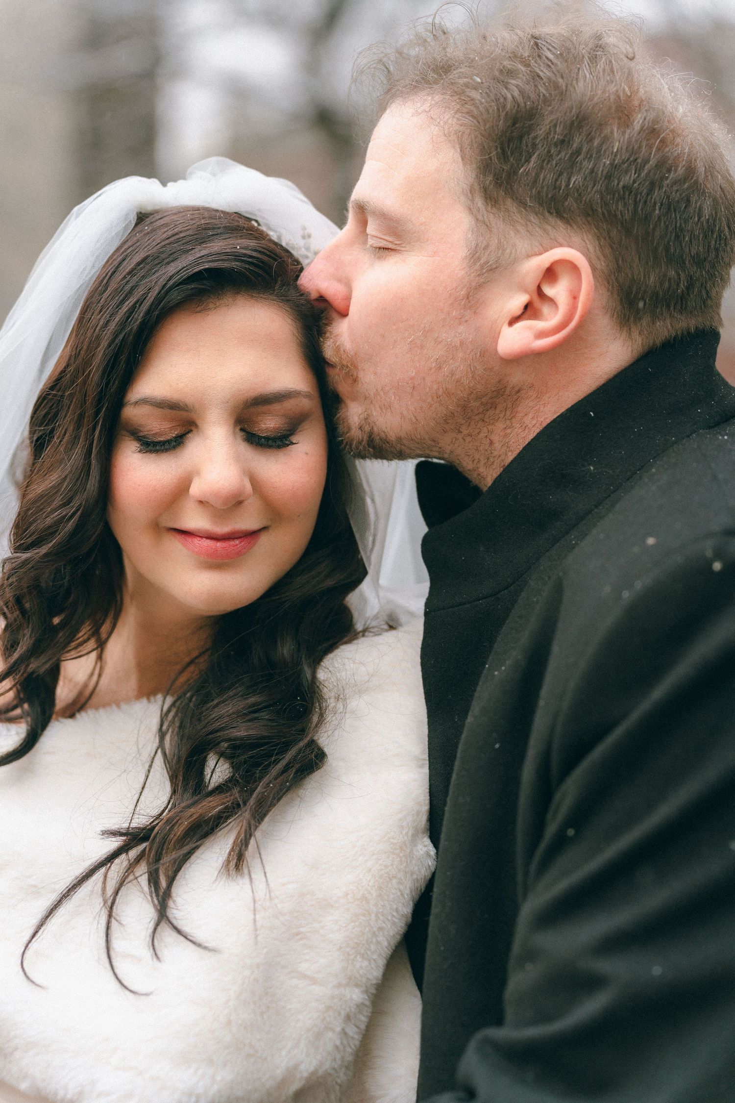 Groom tenderly kisses bride on forehead in romantic winter wedding portrait outdoors.