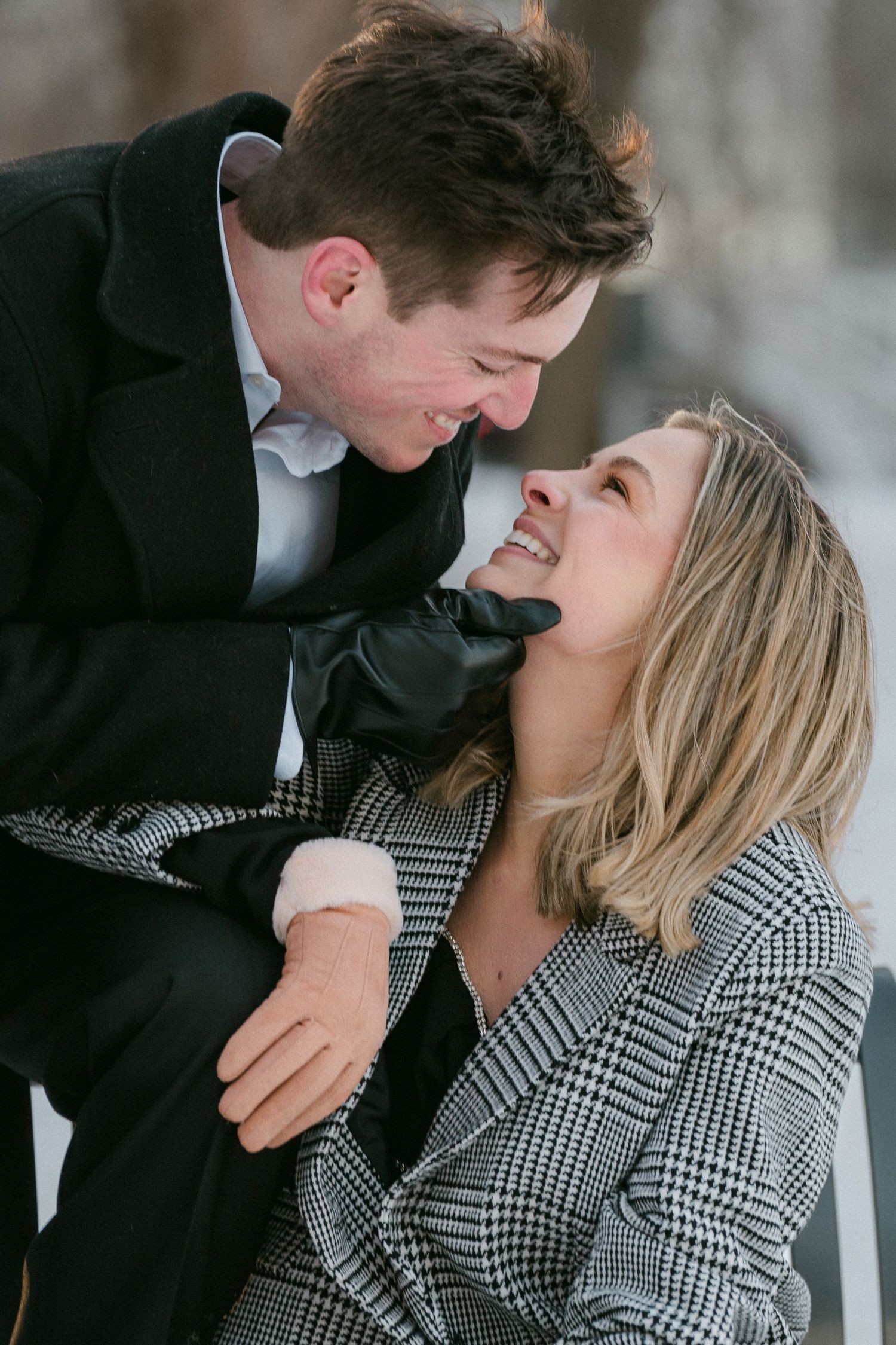 Couple laughing and gazing at each other in snowy winter setting, wearing elegant black and plaid coats.