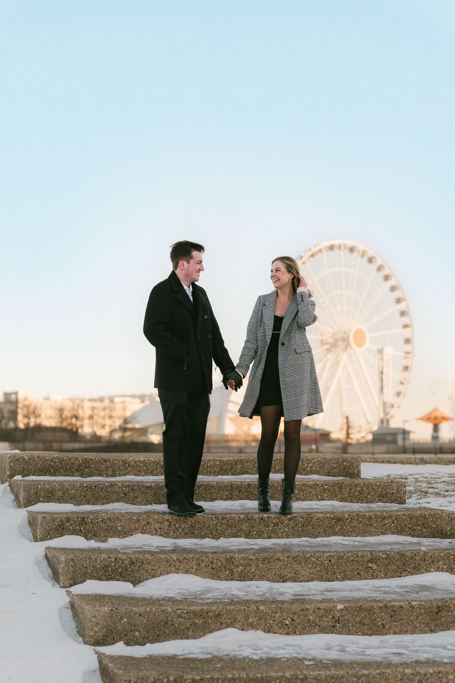 Couple holding hands on snowy stone steps with ferris wheel and golden sunset sky in the background.