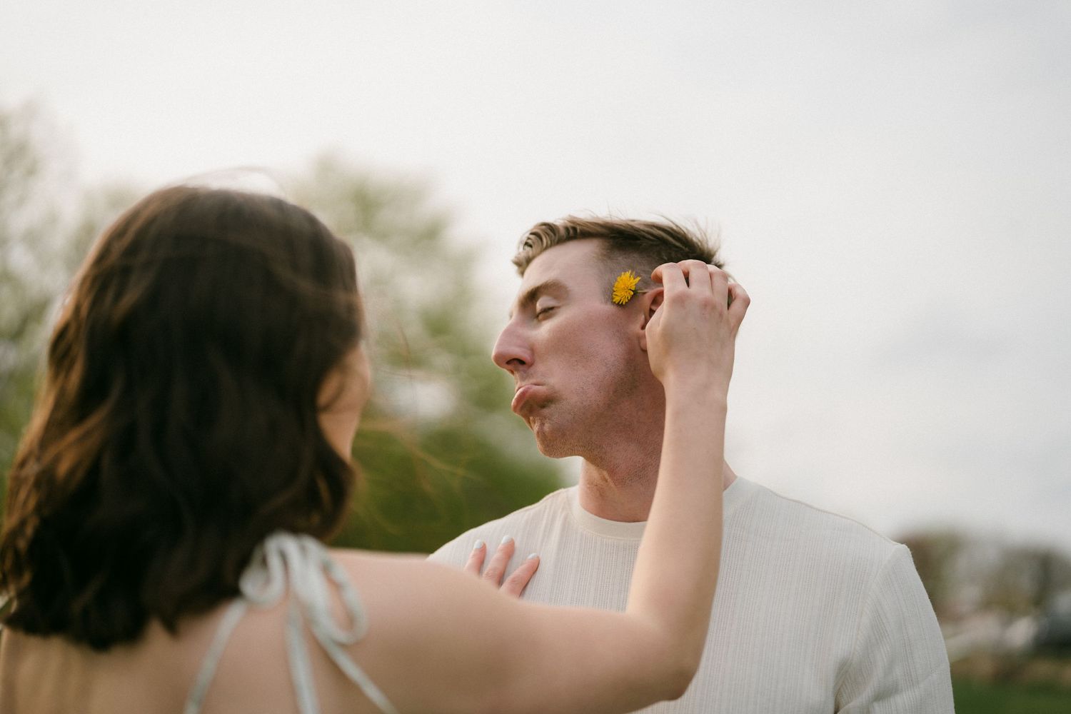 A woman playfully holds a yellow dandelion up to a man's face outdoors.