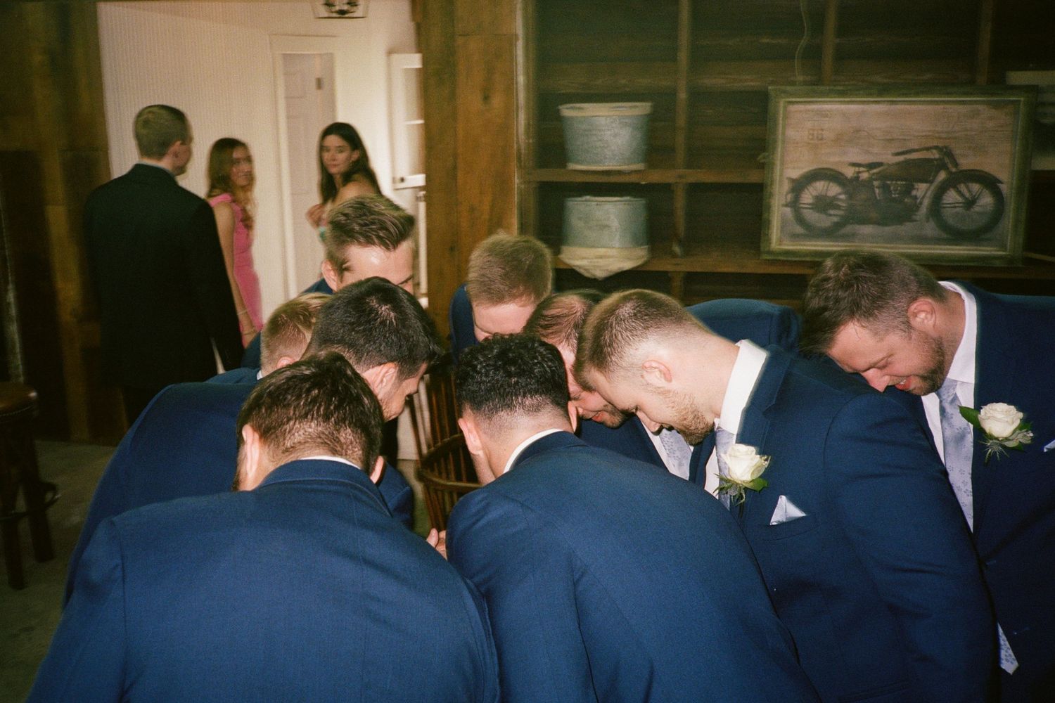 Groomsmen in navy suits huddle together in prayer inside a rustic barn venue before a wedding ceremony.
