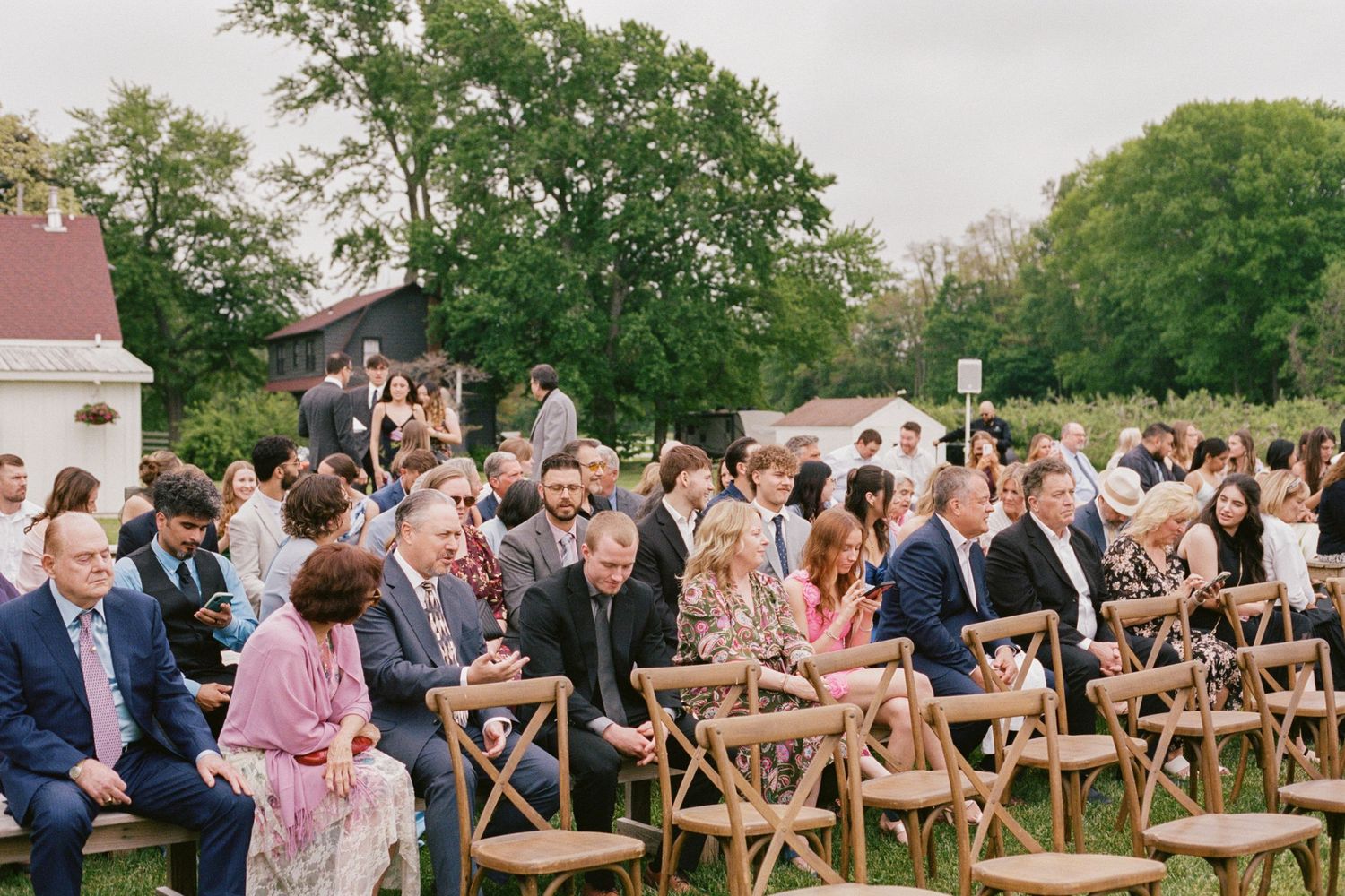 Guests seated in wooden chairs at an outdoor wedding ceremony surrounded by lush green trees.