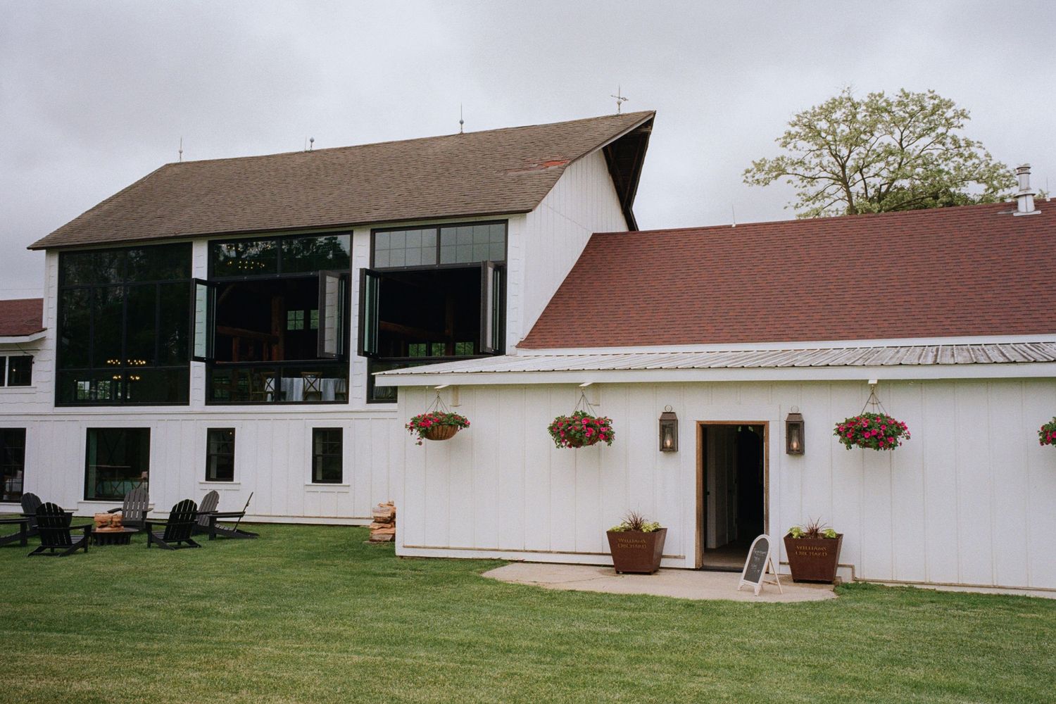 White barn venue with red roof, hanging flower baskets, and modern glass addition surrounded by green lawn.