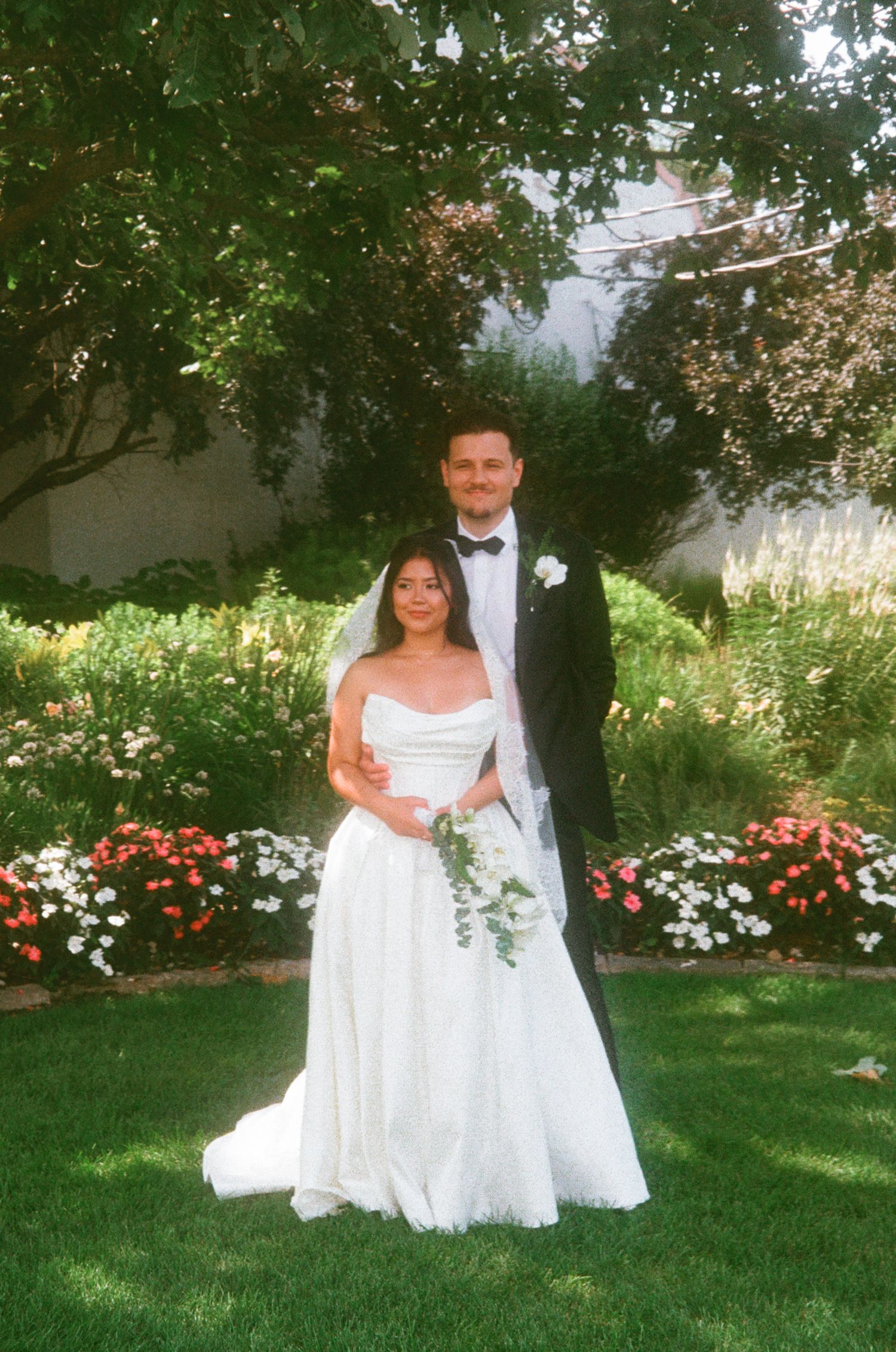 Bride in white strapless gown and groom in black tuxedo pose together in a garden with colorful flowers.