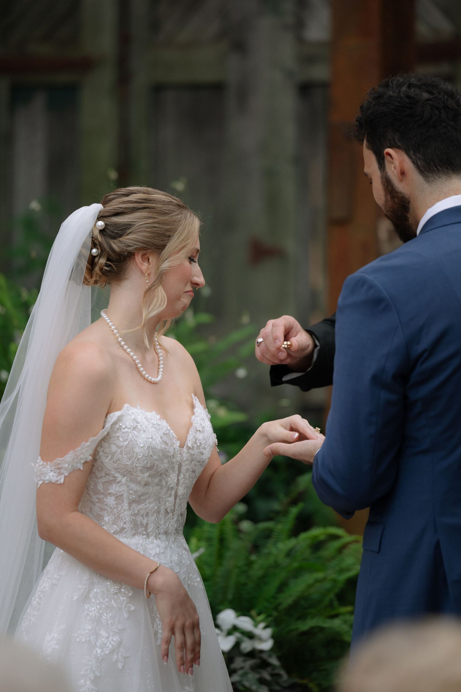 Bride in lace off-shoulder gown and veil receives ring from groom in navy suit during outdoor wedding ceremony.