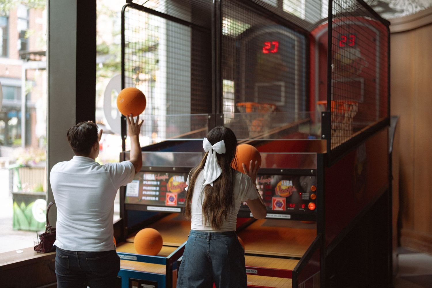 Two people playing arcade basketball game, holding orange basketballs at a colorful arcade machine indoors.