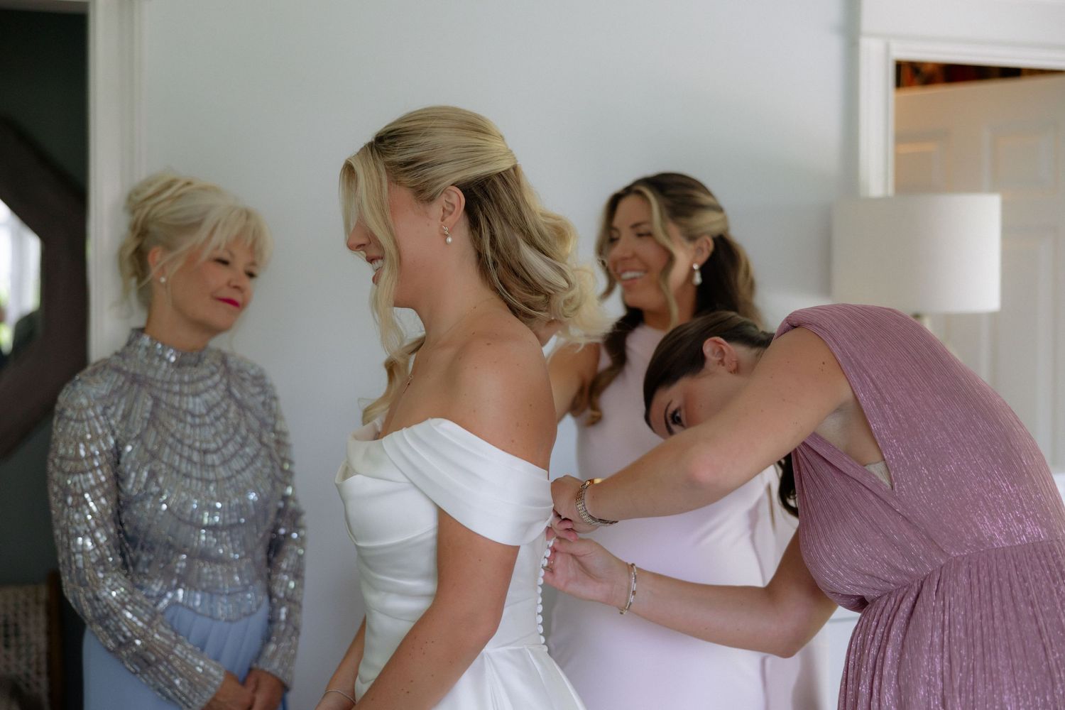 Bridesmaids helping bride button her white off-shoulder wedding gown while guests look on admiringly.