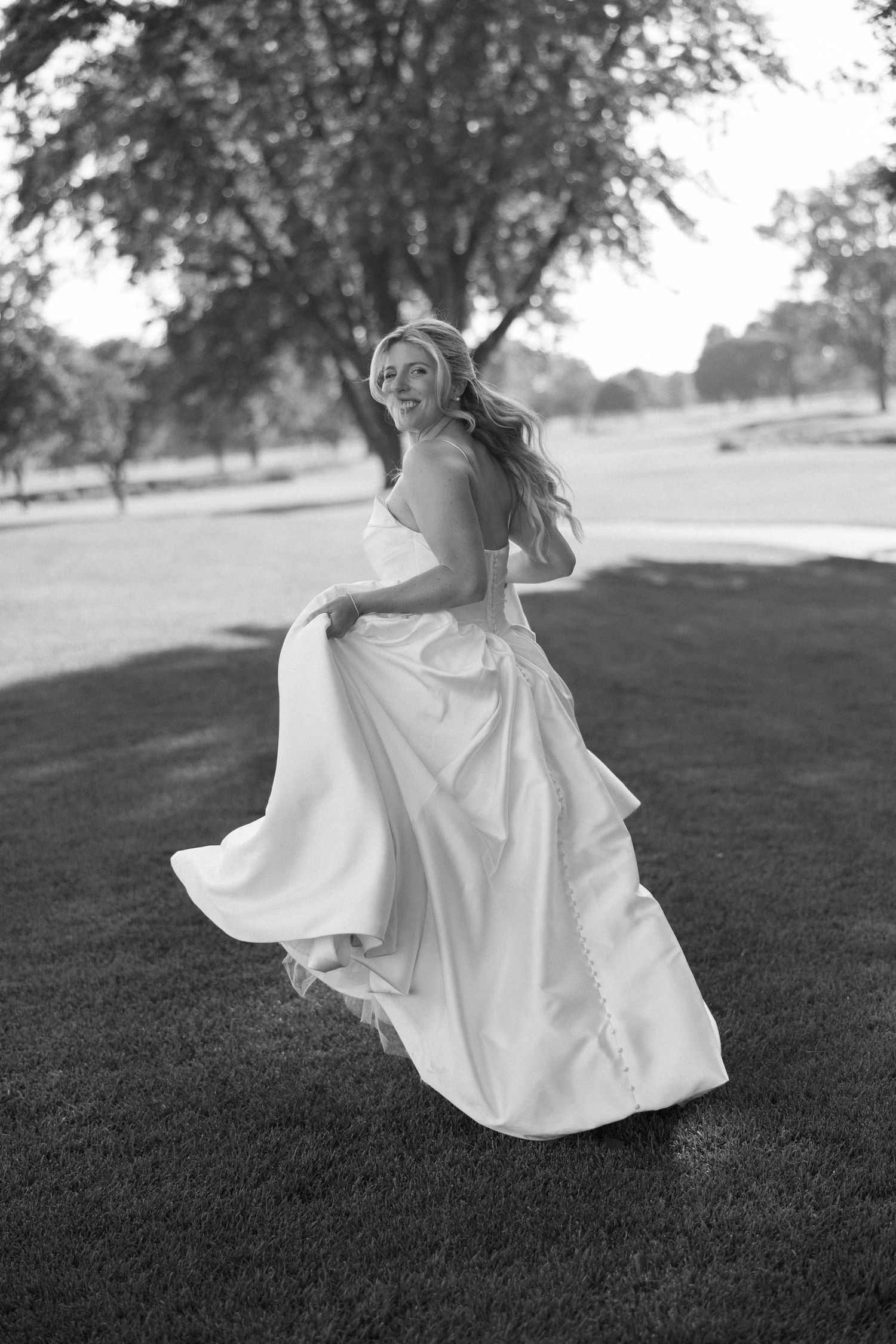 Black and white photo of bride twirling in flowing wedding gown on green lawn with trees.