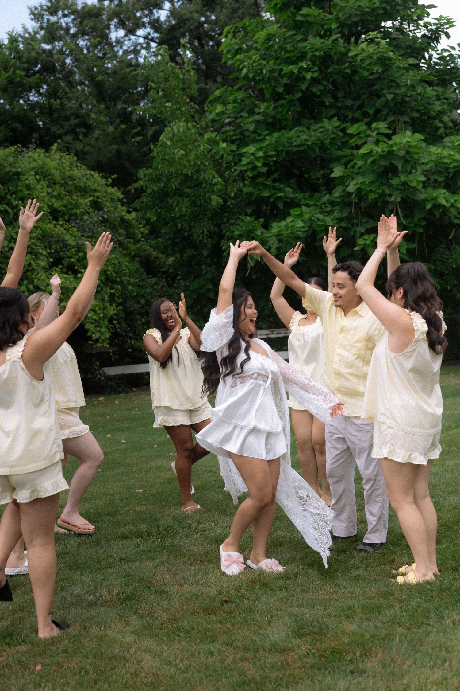 Bride in white dress dancing outdoors with wedding party wearing matching yellow outfits on green lawn.