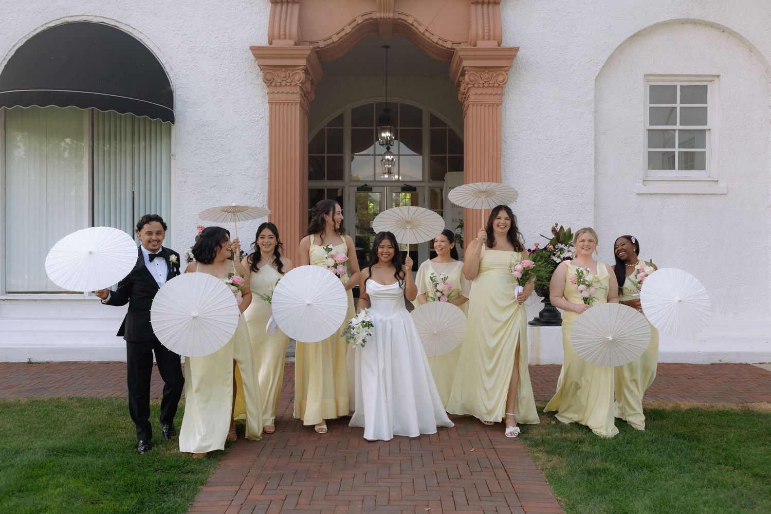 Wedding party poses outside white building, bride in white gown with bridesmaids in yellow dresses holding white parasols.