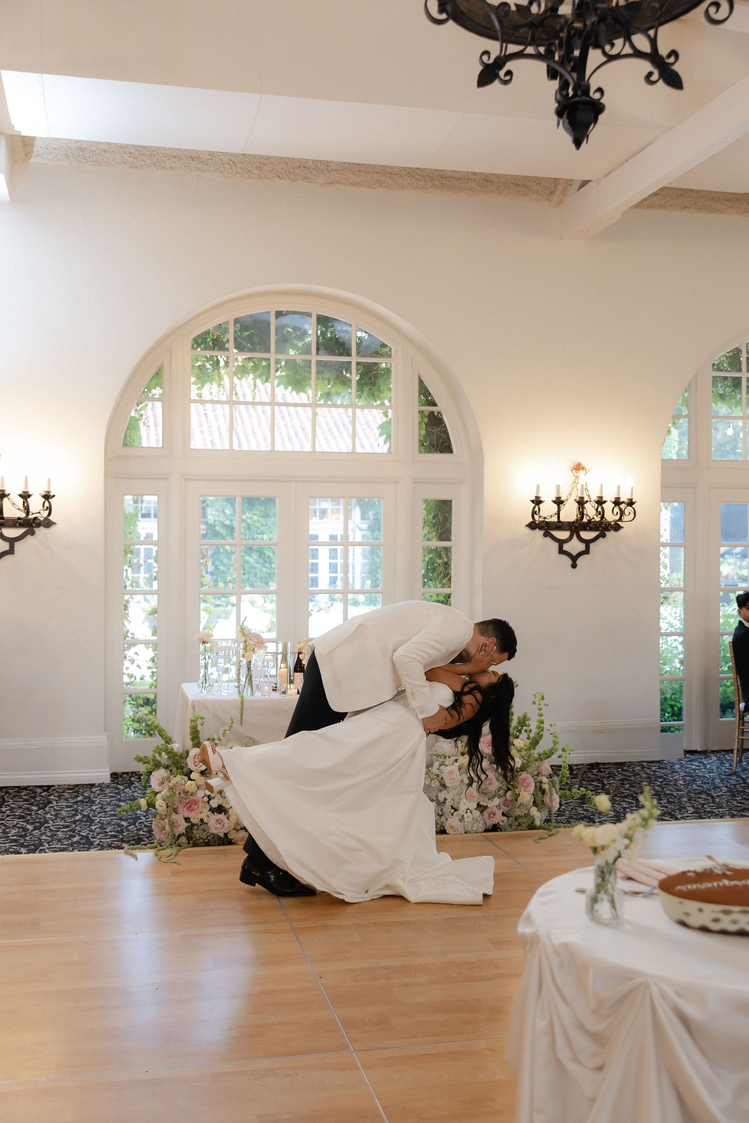 Groom dipping bride during first dance in elegant white ballroom with arched windows and chandelier.