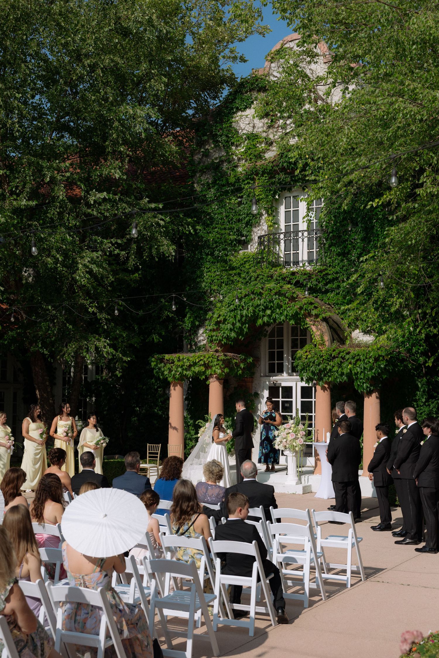 Outdoor wedding ceremony with guests seated in white chairs before an ivy-covered stone mansion.