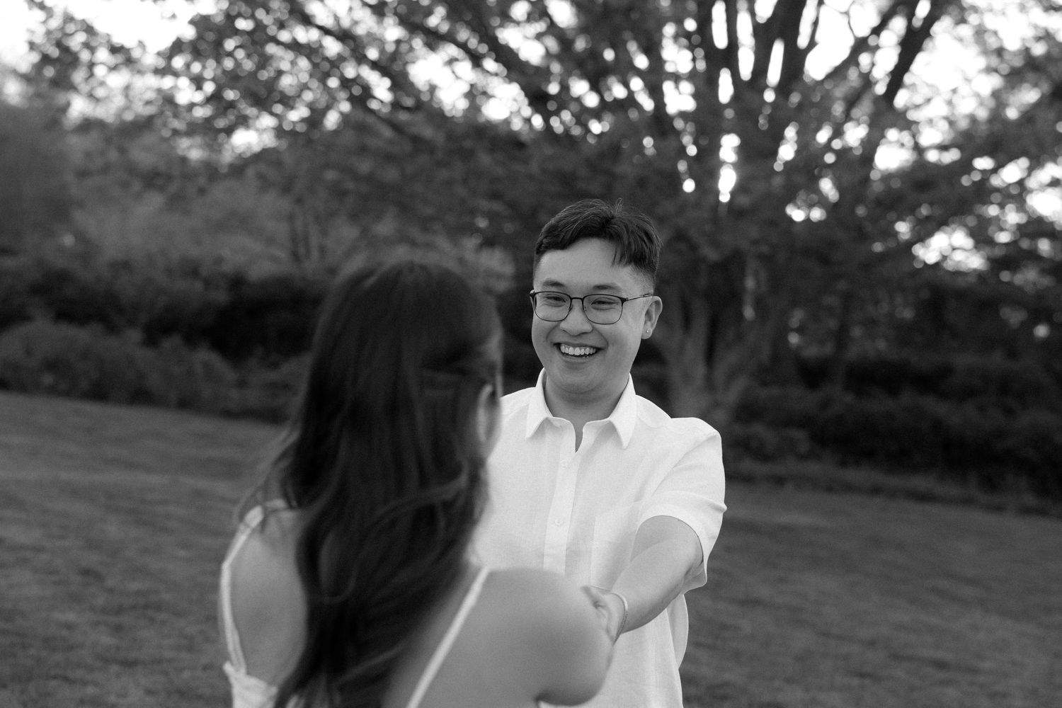 Black and white photo of a smiling man in white shirt reaching out to a woman outdoors in a park setting.