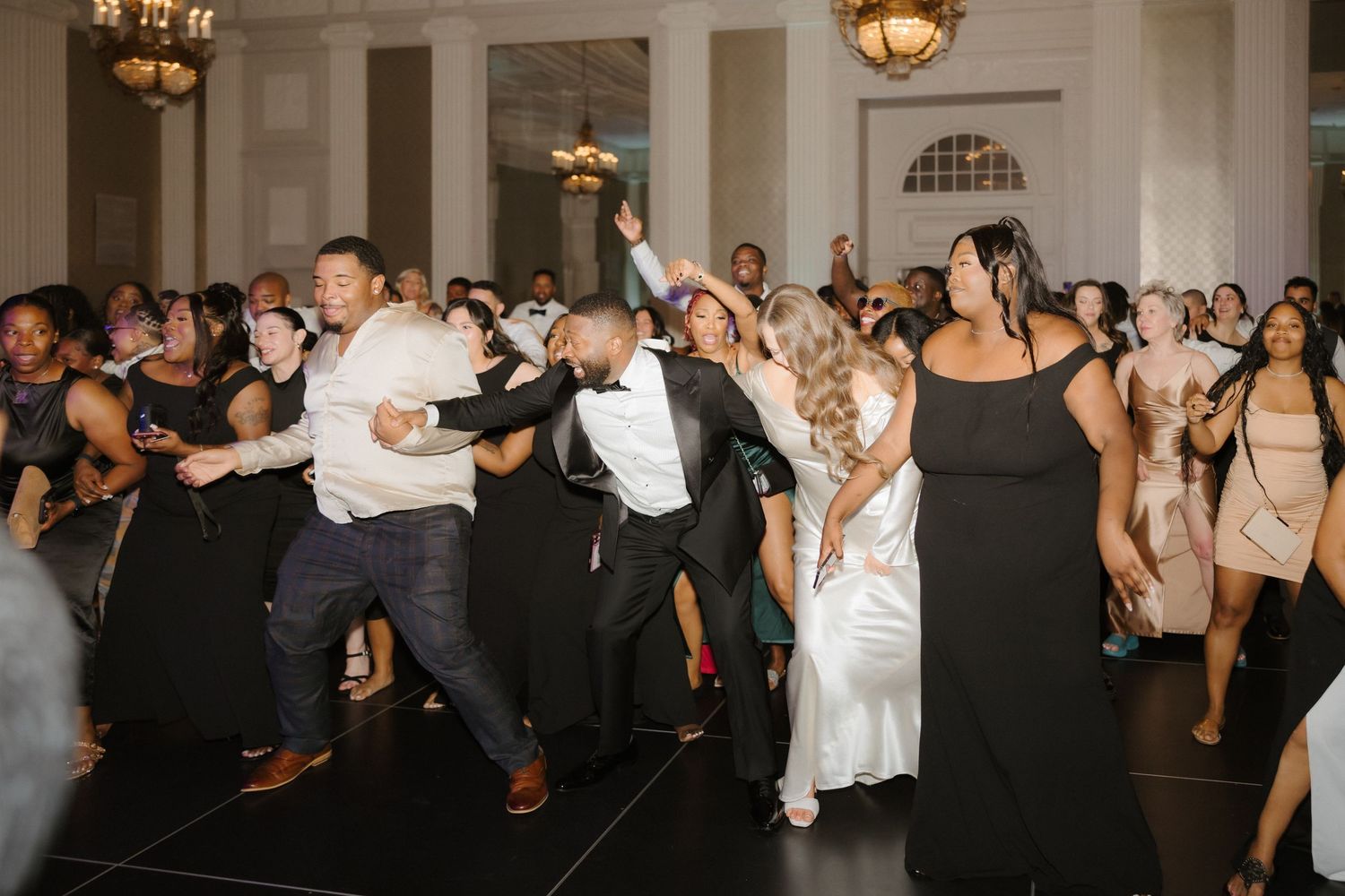 Wedding guests dancing joyfully at elegant reception venue, bride in white gown celebrating with friends on dance floor.