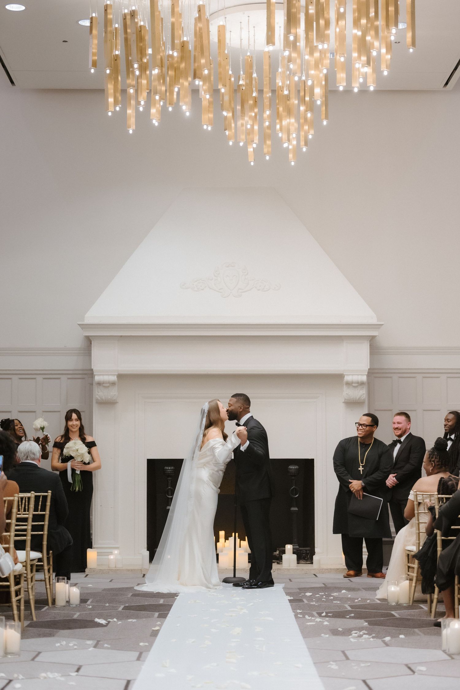 Bride and groom share first kiss at elegant indoor wedding ceremony with gold chandelier and white fireplace backdrop.