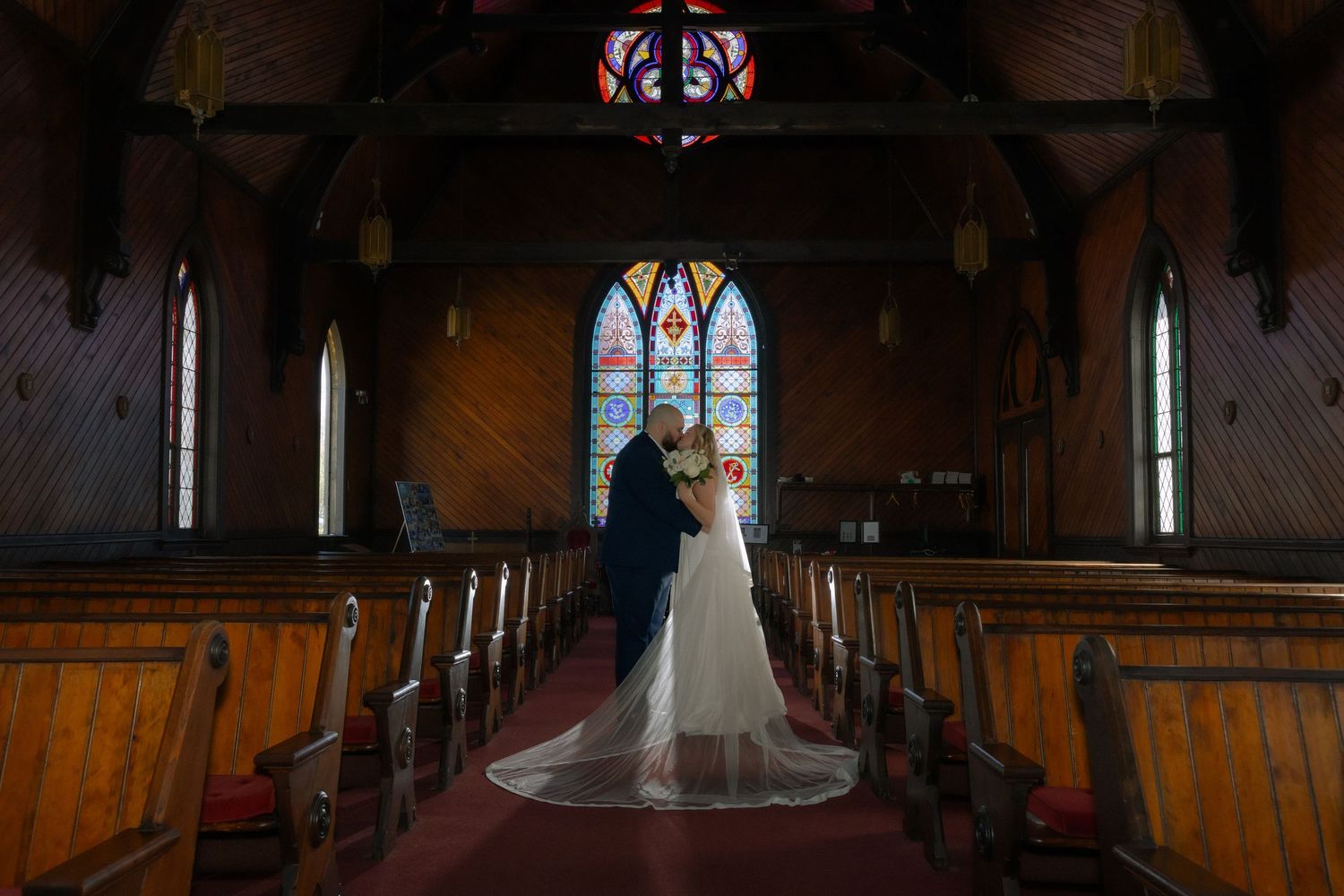 Bride and groom stand at altar in historic wooden church with stunning stained glass windows during wedding ceremony.