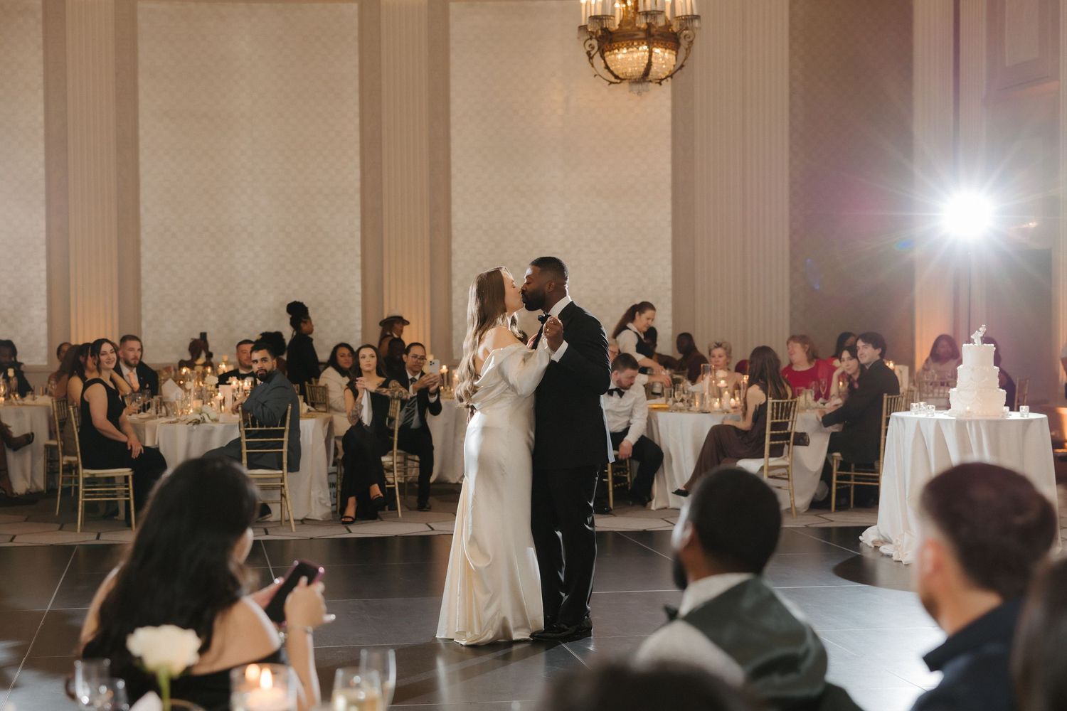 Bride and groom share their first dance at an elegant wedding reception in a grand ballroom with guests seated at candlelit tables.