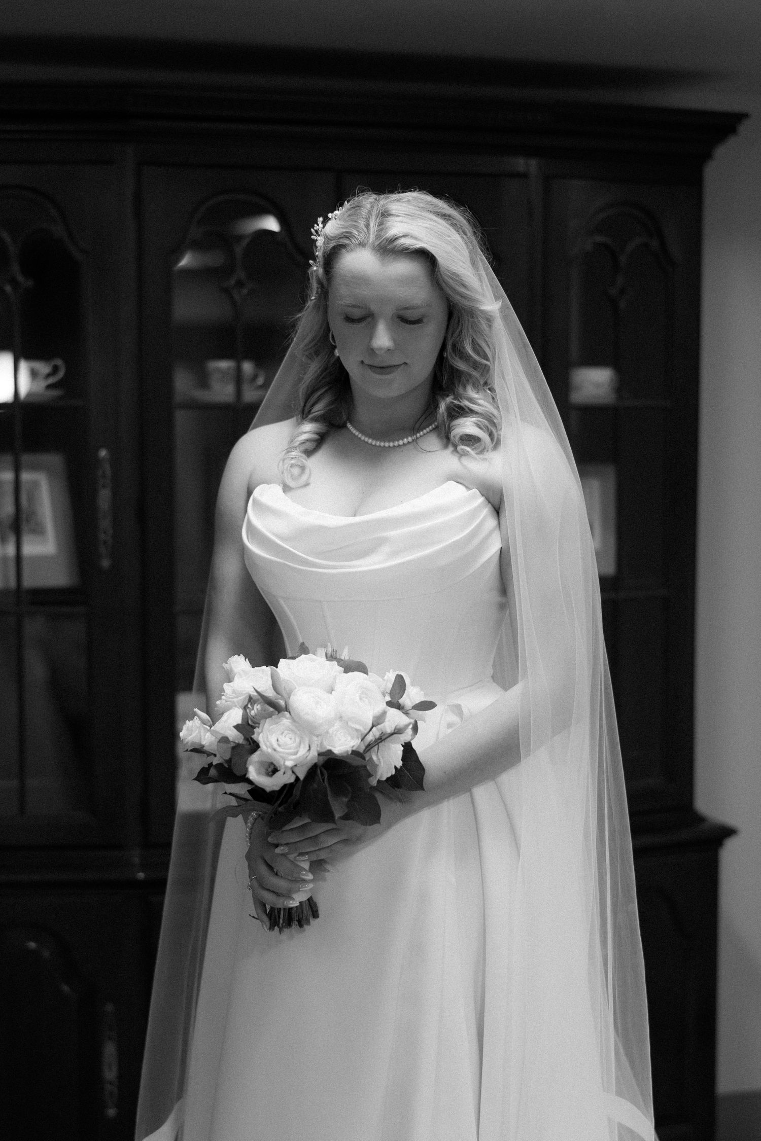 Black and white photo of a bride in a strapless gown and veil holding a bouquet of roses, looking down gracefully.