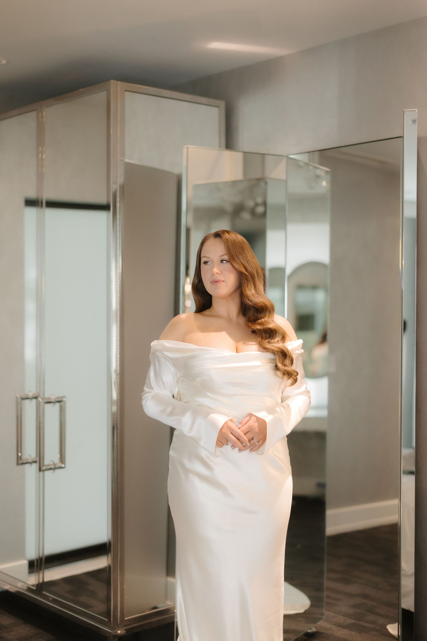 Bride in elegant off-shoulder white satin gown posing in a mirrored bridal boutique fitting room.