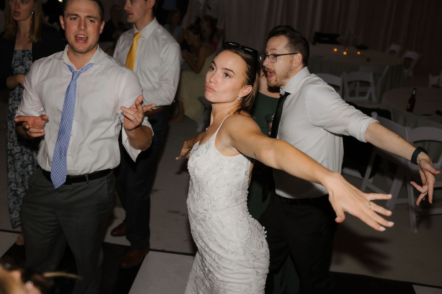 Bride in lace wedding dress dancing joyfully with guests at an indoor wedding reception.